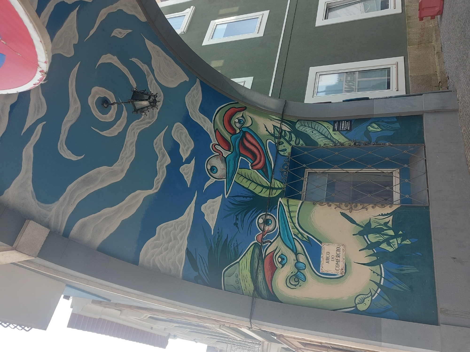 The image shows a street view in Aveiro, Portugal, with a building facade decorated with a large mural. The mural features two stylized birds with prominent eyes and red and blue markings, surrounded by green foliage. Above the birds, the mural depicts a blue sky with swirling white clouds and a vintage-style street lamp. A sign reading "Arco do Comércio" is integrated into the mural design. Above the mural, there are windows of a green building. A road sign is also visible.