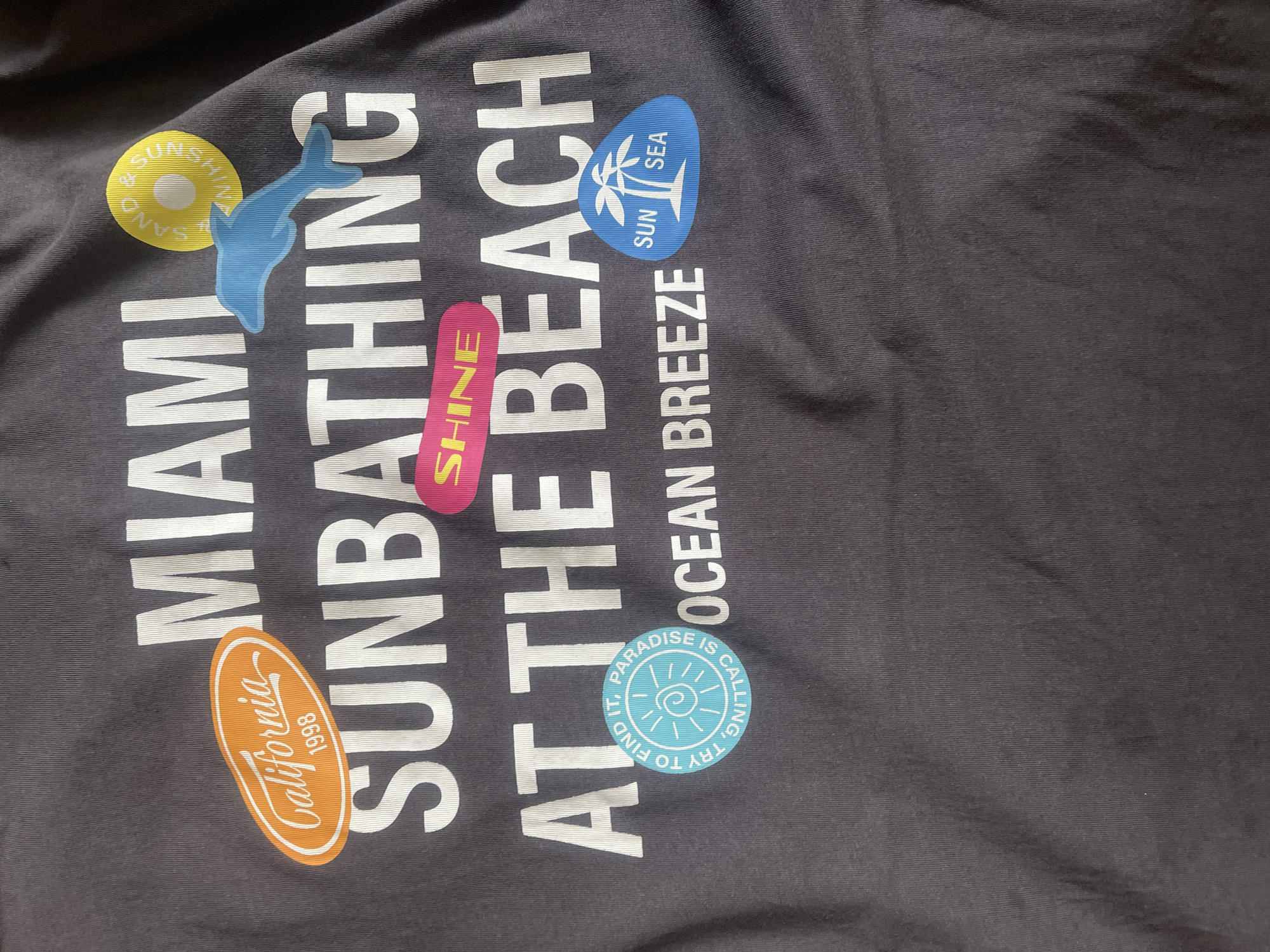 The image shows a close-up view of a dark gray t-shirt with white text and colorful graphic elements. The text on the t-shirt reads "MIAMI SUNBATHING AT THE BEACH OCEAN BREEZE".
Scattered among the text are several graphic elements:
* A yellow circle with the text "& SUNSHINE & SAND" written in a wavy font.
* A blue dolphin shape.
* A pink rectangle with the word "SHINE" written in yellow.
* A blue oval shape with a palm tree inside and the words "SUN" and "SEA".
* An orange oval shape with the text "California 1998".
* A teal-colored circle with a sun graphic and the text "PARADISE IS CALLING, TRY TO FIND IT."
The t-shirt is laid flat and appears to be slightly wrinkled. The image is taken from a top-down perspective.