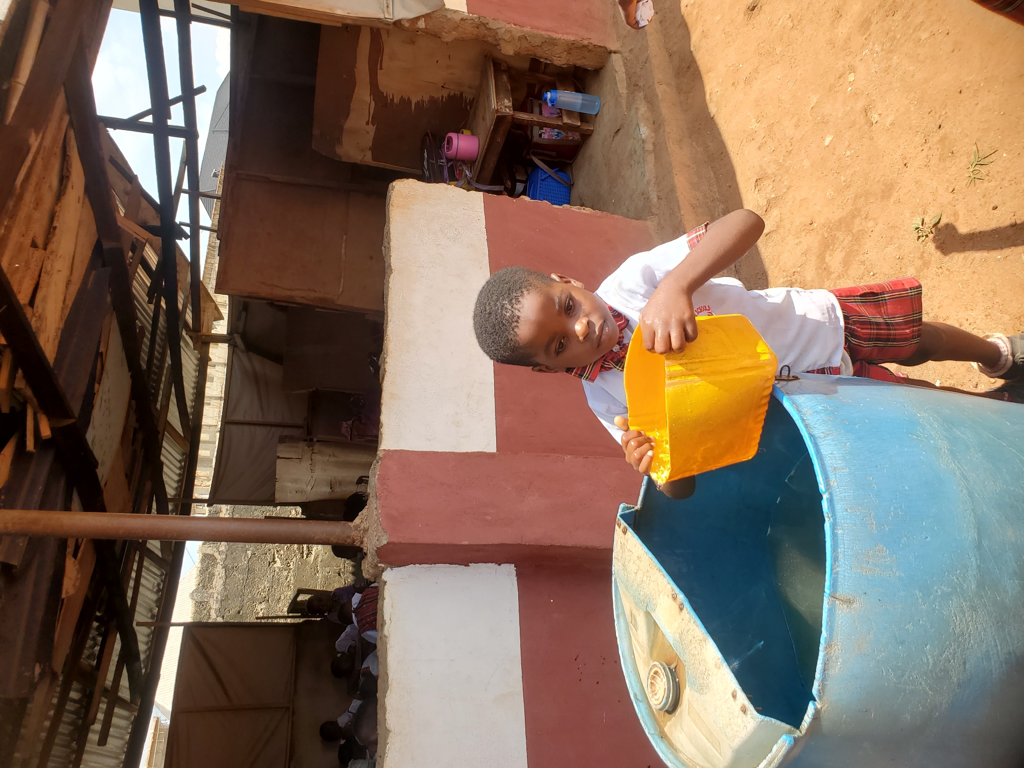 In Ukwu-Abwa, Nigeria, a child in a white shirt, plaid shorts, and a bow tie pours a yellow container into a blue barrel. The setting appears to be outdoors near a structure with brick and wood.