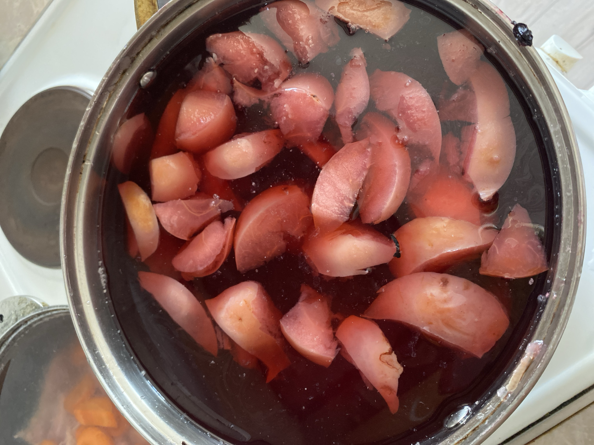 A close-up, overhead shot shows sliced, pinkish-red fruit, possibly plums or peaches, cooking in a dark, reddish liquid within a silver pot. The pot is on a white stovetop. Bubbles are visible in the liquid, indicating it is simmering. The fruit pieces are varied in size and shape, with some showing the skin and others appearing softer and more cooked. The pot has a metallic rim and a silver handle is partially visible on the right side. In the background, another pot containing orange slices is partially visible, also appearing to be on the stovetop. The overall environment suggests a kitchen setting. The image does not contain any identifiable landmarks or context that would allow for its specific location within the city of Chornomorsk, Ukraine.