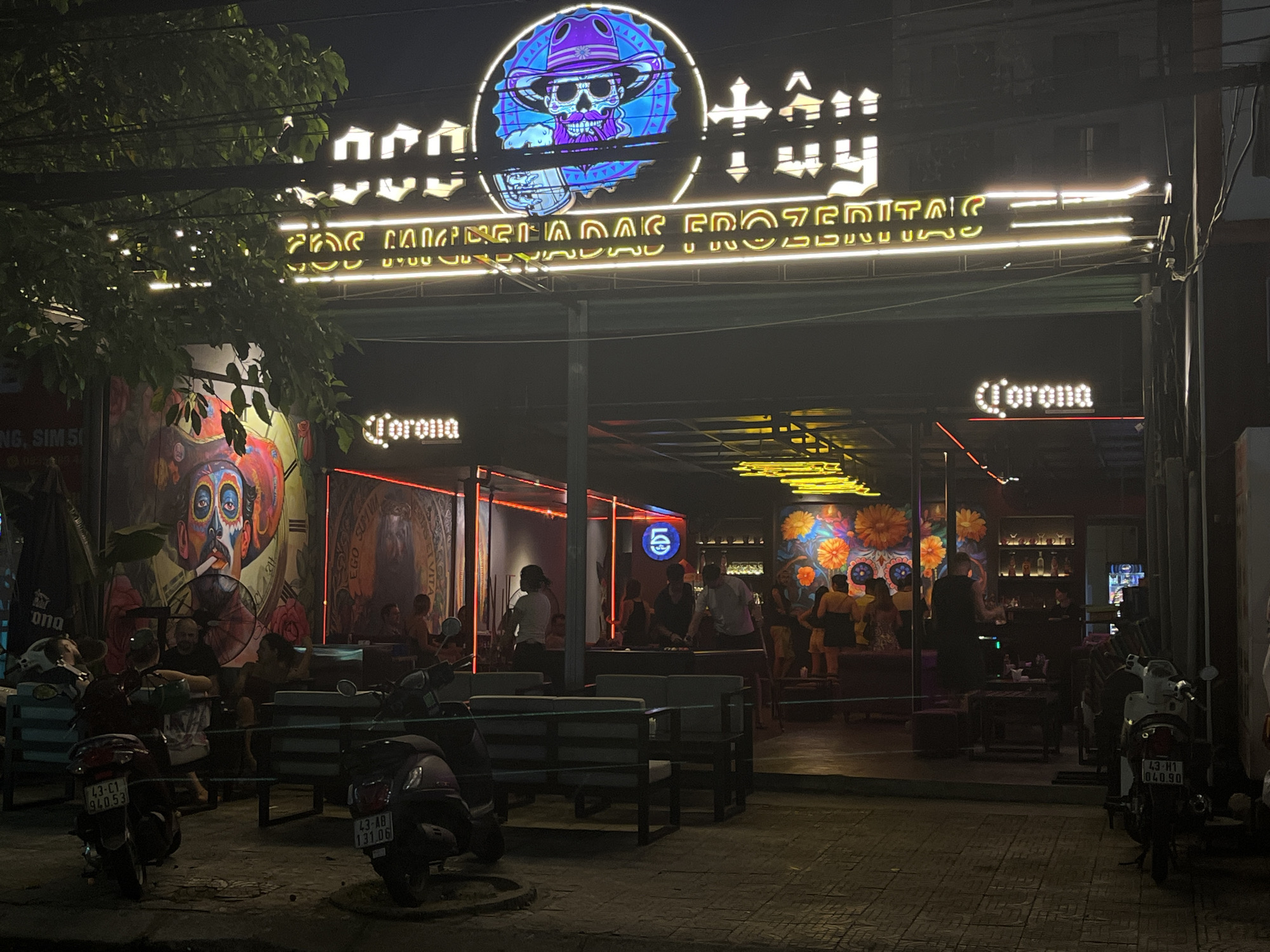 The image shows the exterior of a bar/restaurant in Đà Nẵng, Vietnam at night. The facade is open, with several people inside, seated at tables. On the street, motorbikes are parked and some individuals are sitting on benches. The establishment has bright, illuminated signage above the entrance, featuring the name "Tây Hy" with a skull graphic, and another sign that reads "LAS MICHELADAS FROZENITAS". "Corona" signs are also displayed. The overall environment is dark, illuminated by the lights from the establishment and surrounding street lights.