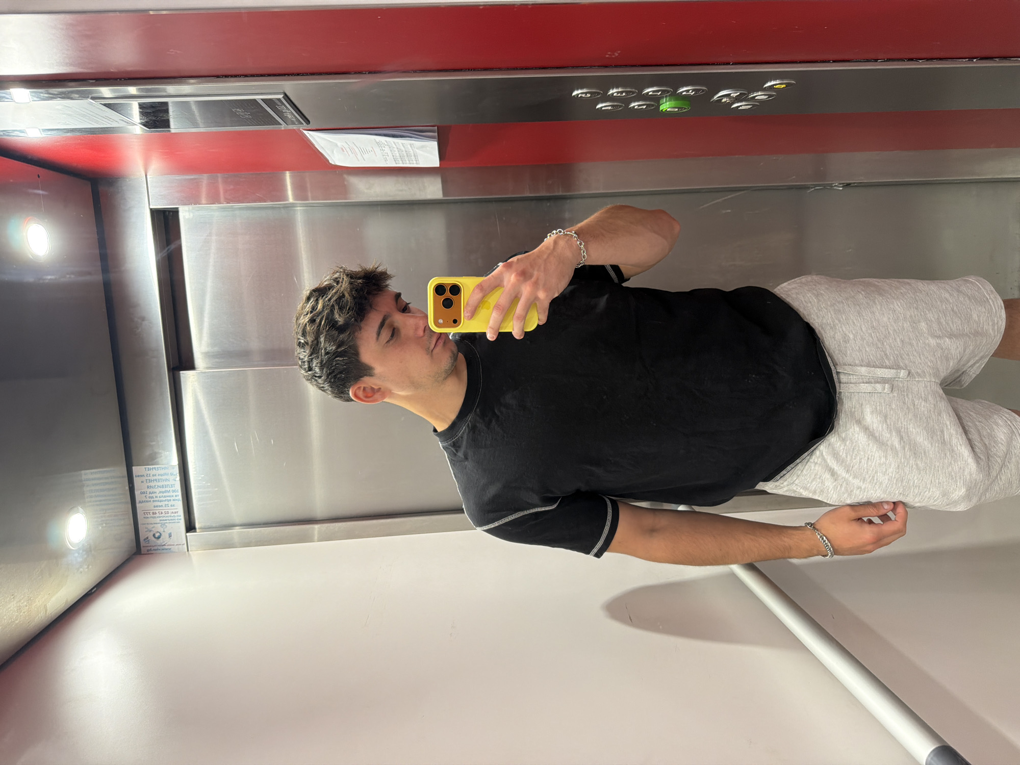 A young man is taking a selfie in an elevator. He is holding a yellow phone in his right hand. He is wearing a black t-shirt, grey shorts, and a silver bracelet. The elevator has red and silver walls, along with control buttons. The location may be in Sofia, Bulgaria.