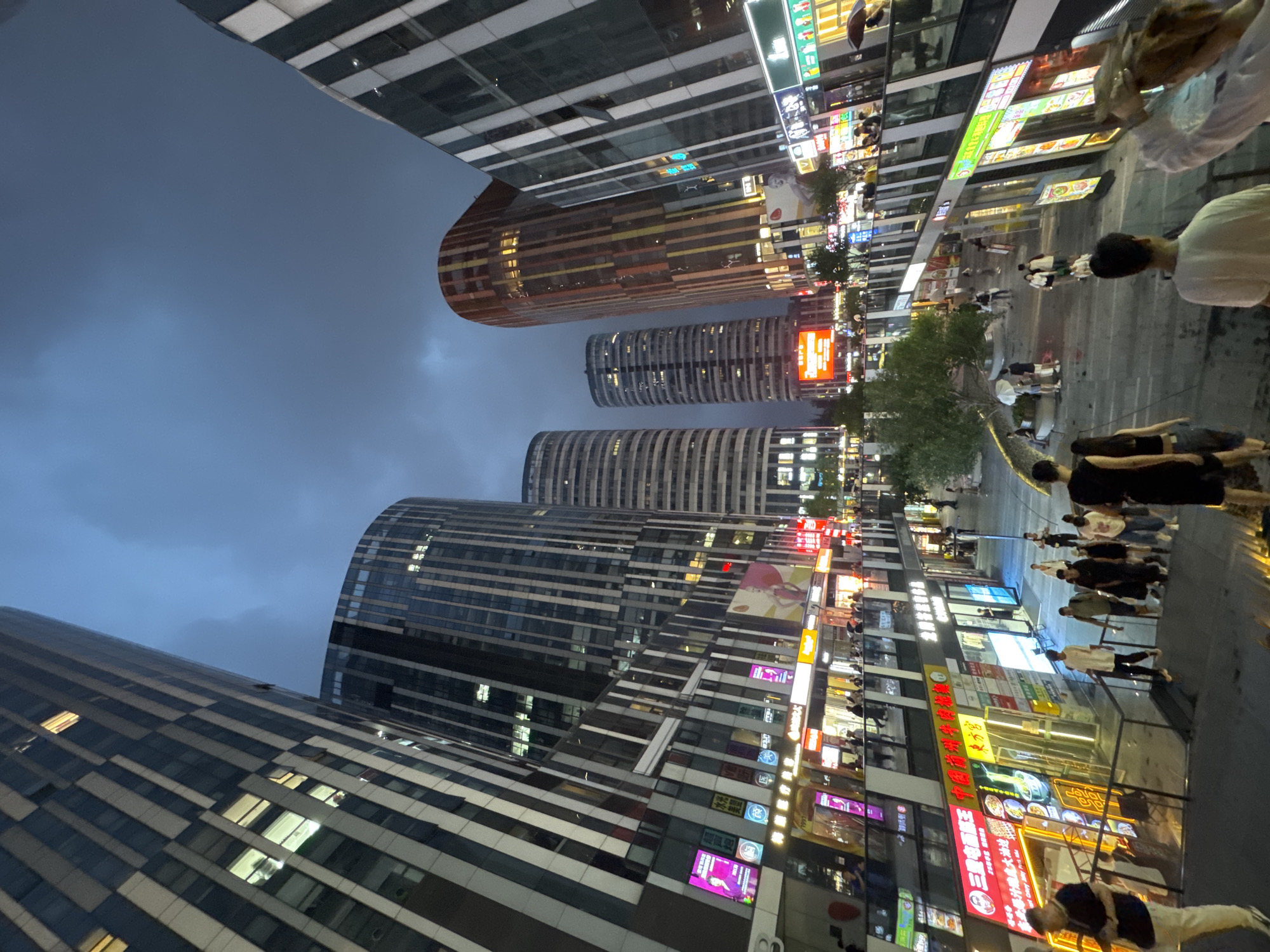 The image depicts a bustling pedestrian area in Chaoyang District, China, at dusk or early evening. Towering modern skyscrapers with reflective glass facades dominate the background, some illuminated from within. The foreground shows a multi-level plaza filled with people walking, socializing, and patronizing various shops and restaurants. Brightly lit signage in Chinese characters is prominent, advertising businesses such as restaurants, clothing stores, and possibly electronics shops. The atmosphere appears lively and active, with individuals and groups scattered throughout the scene. Some people are dressed casually, while others are in more formal attire, suggesting a diverse range of activities and purposes for being in the area. The ground level appears to be a walkway or street, with elevated walkways and stairs connecting different levels of the complex. A few trees are visible, adding a touch of greenery to the urban landscape. The sky is a deep, overcast blue, indicating potentially inclement weather or simply the transition into night.