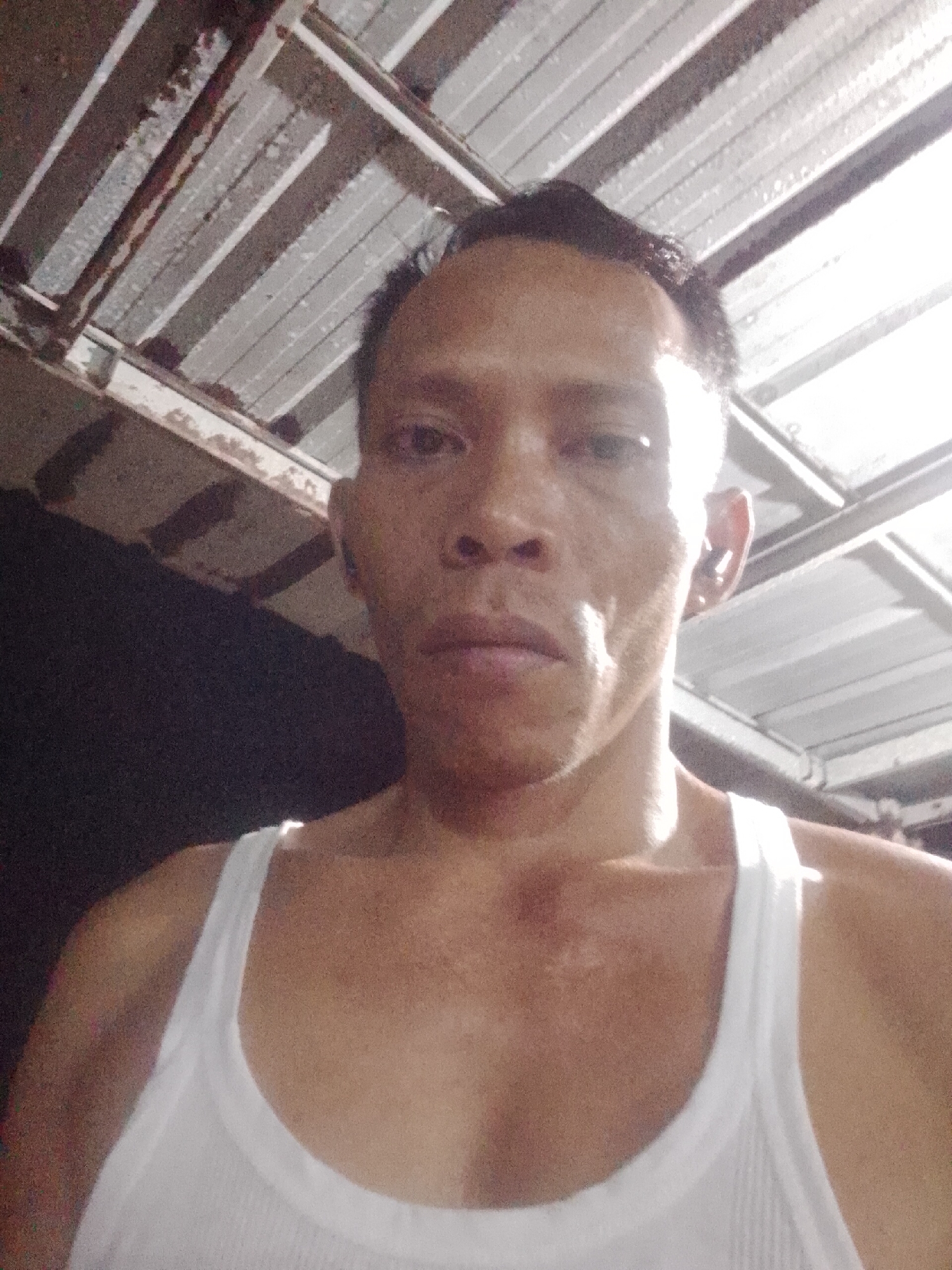 A close-up, self-portrait photograph shows a person in Bukit Ketok, Indonesia. The individual has short, dark hair, and a neutral facial expression. They are wearing a white tank top. The lighting is uneven, with strong highlights on the right side of the face and upper chest. The background consists of a corrugated metal roof structure, partly in shadow. The setting appears to be an outdoor or semi-outdoor space.