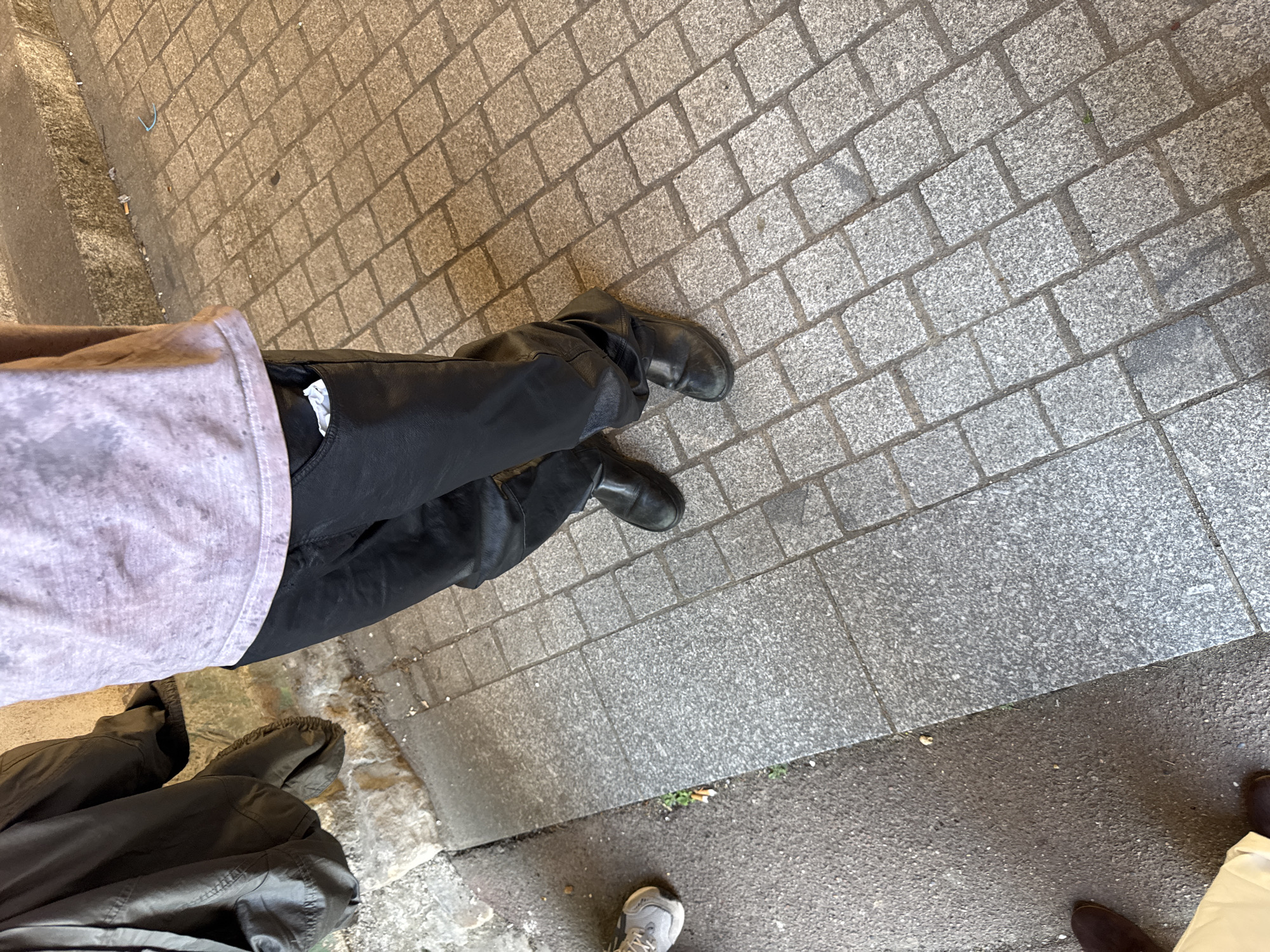 A view from above shows a person standing on a grey cobblestone street. The person is wearing black pants and black boots. The legs of another person in black pants and dark boots are partially visible in the lower left corner. Also visible are other legs with a brown shoe and a grey sneaker. The location appears to be an urban setting, potentially a sidewalk in Palaiseau, France.