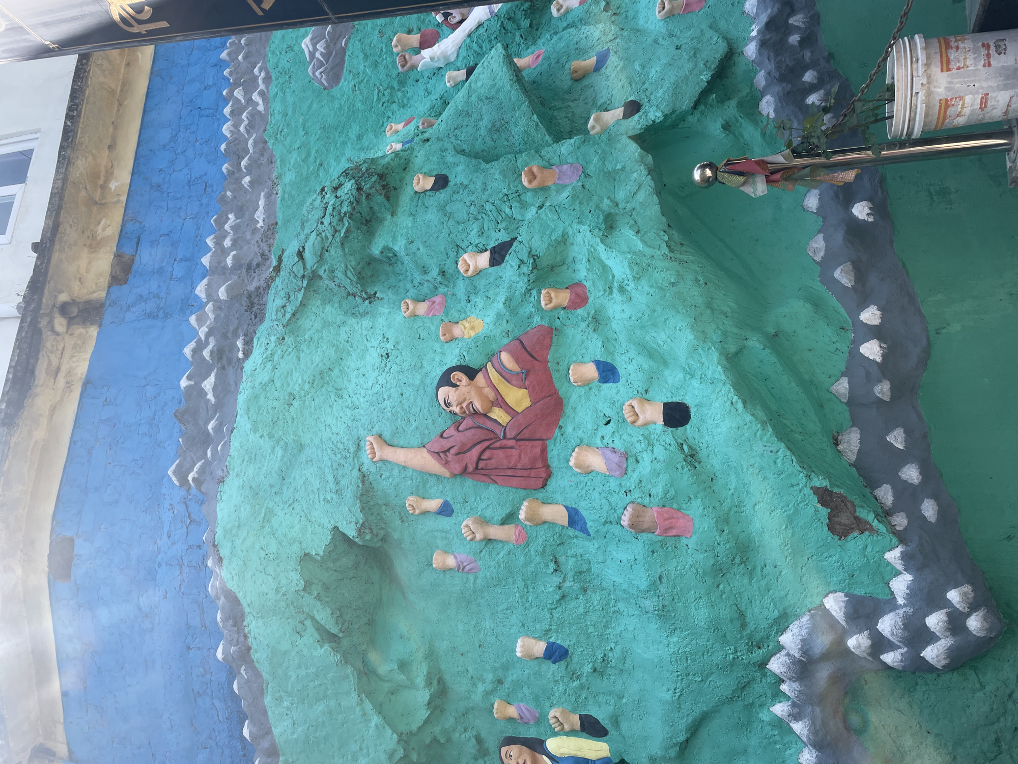 The image shows a climbing wall mural in Dharamshala, India. The wall is painted a light green color with gray areas. Figures of people are depicted climbing the wall, using sculpted handholds of various colors. The central figure is wearing a red and yellow robe and is reaching for a hold. The wall also has an uneven, textured surface. A blue wall is visible to the left. A metal pole and container are visible on the upper right. A building is seen in the upper left.