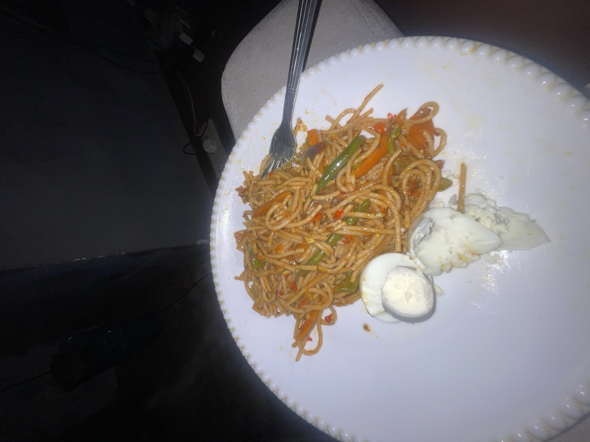 A plate of spaghetti with vegetables and sliced boiled eggs is on a white decorative plate. A silver fork is partially visible, resting on the spaghetti. The scene is dimly lit and the surrounding area appears to be dark, though a portion of a chair or couch is visible behind the plate. There is no indication of location in Kurunduma, Nigeria or any other identifying information in the image.