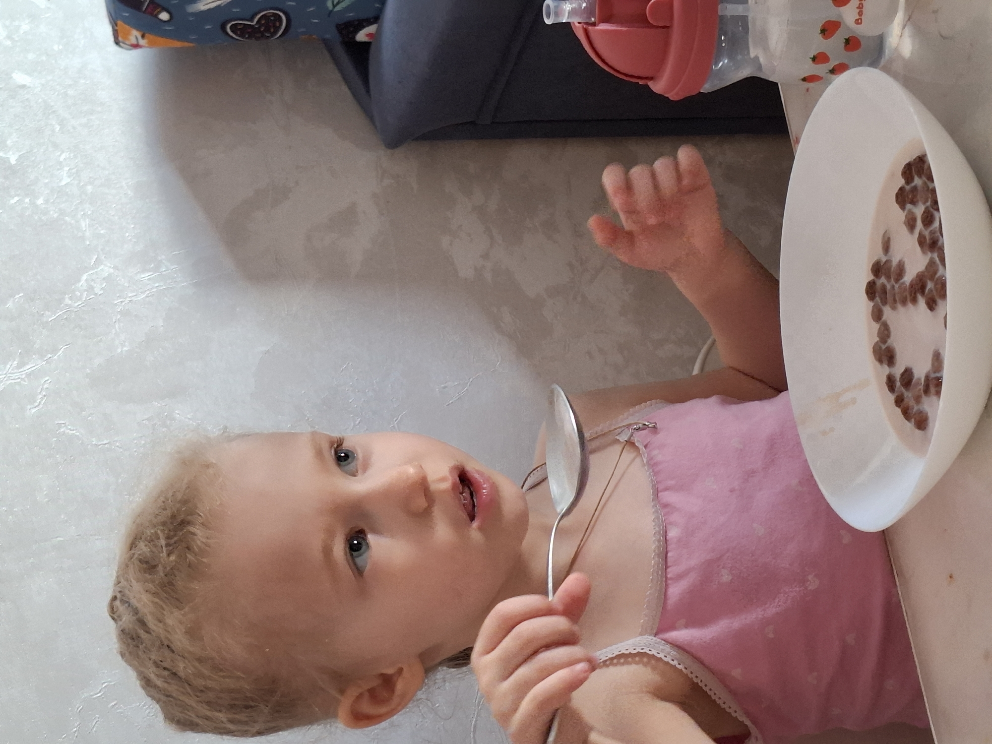 The image captures a young child, likely a girl, with fair skin, light blonde hair, and blue eyes, engaged in a mealtime activity. She is wearing a pink sleeveless top with a delicate lace trim along the edges. The child holds a silver metal spoon in her right hand, positioned near her mouth, suggesting she is either eating or about to eat.
In front of her, on what appears to be a light-colored table or countertop, is a white bowl filled with chocolate-flavored cereal puffs submerged in milk. To the right of the bowl, a clear sippy cup with a pink lid and straw is visible. The sippy cup is decorated with small red strawberry illustrations, and the word "Baby" can be faintly seen printed on its side.
The child is looking slightly upwards and to her left, with a neutral and observant expression, possibly at an adult or another person out of frame. In the background, part of a dark grey object, possibly a chair or sofa cushion, is visible. This object is covered with a blue fabric patterned with various cartoon-like designs, including foxes and hearts. The setting appears to be an indoor environment, likely a home dining area. This scene takes place in Chaikovsky, Russia.