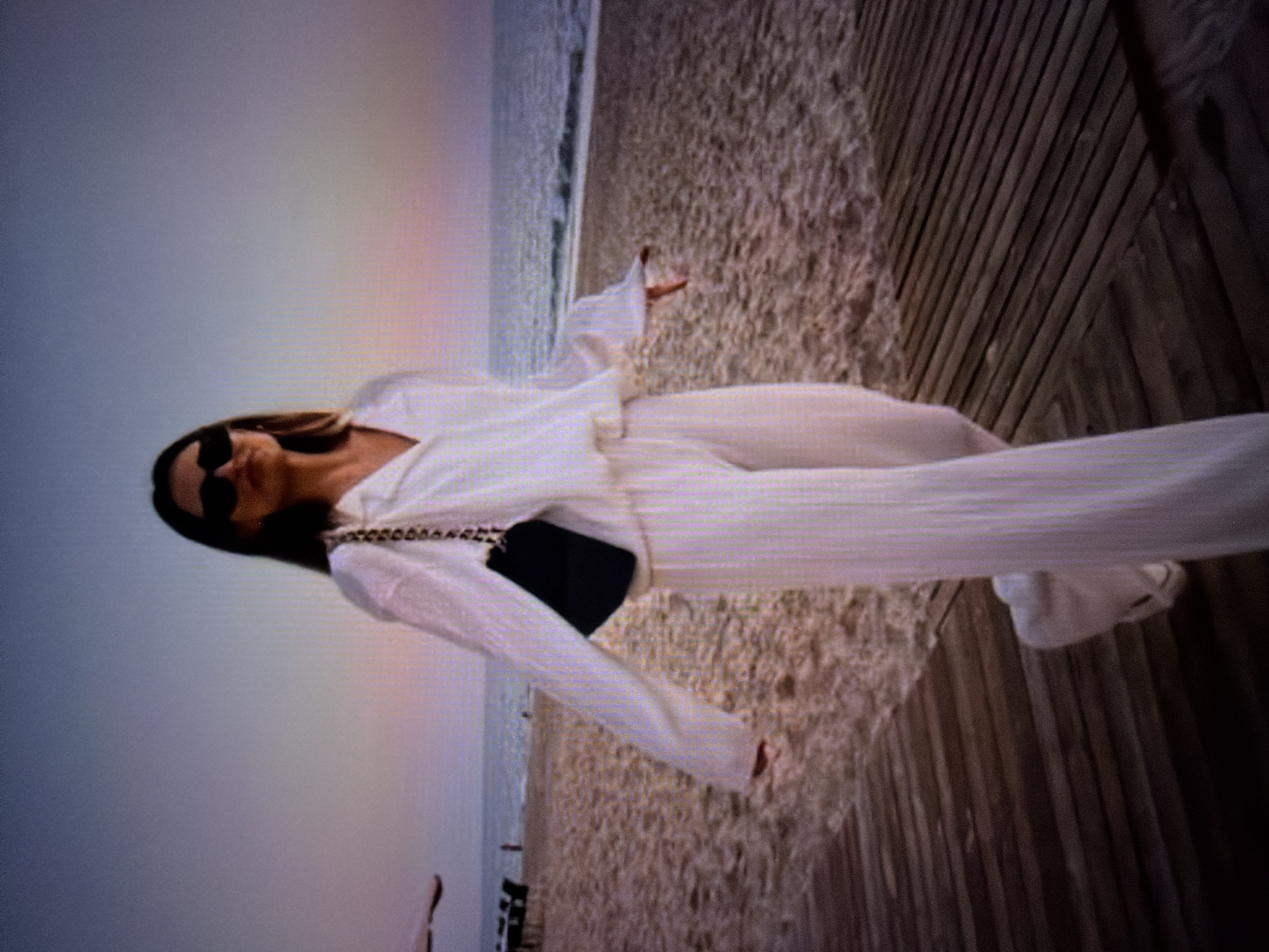 The image shows a person dressed in a white outfit, possibly a matching set of pants and a long-sleeved shirt, standing on a wooden boardwalk. The boardwalk is situated next to a sandy beach, with the ocean visible in the background. The sky is a pale, muted grey, suggesting it might be early morning, late evening, or an overcast day. The person is wearing sunglasses and a dark clutch bag. Their posture suggests they are posing or perhaps enjoying the view. The overall mood is calm and serene, possibly a moment of leisure or vacation. The wooden planks of the boardwalk are arranged diagonally, creating a visual pattern. The sand appears light in color, and the ocean has a gentle surf. The lighting is soft, not casting harsh shadows, further supporting the idea of diffused daylight. No visible text is present in the image.