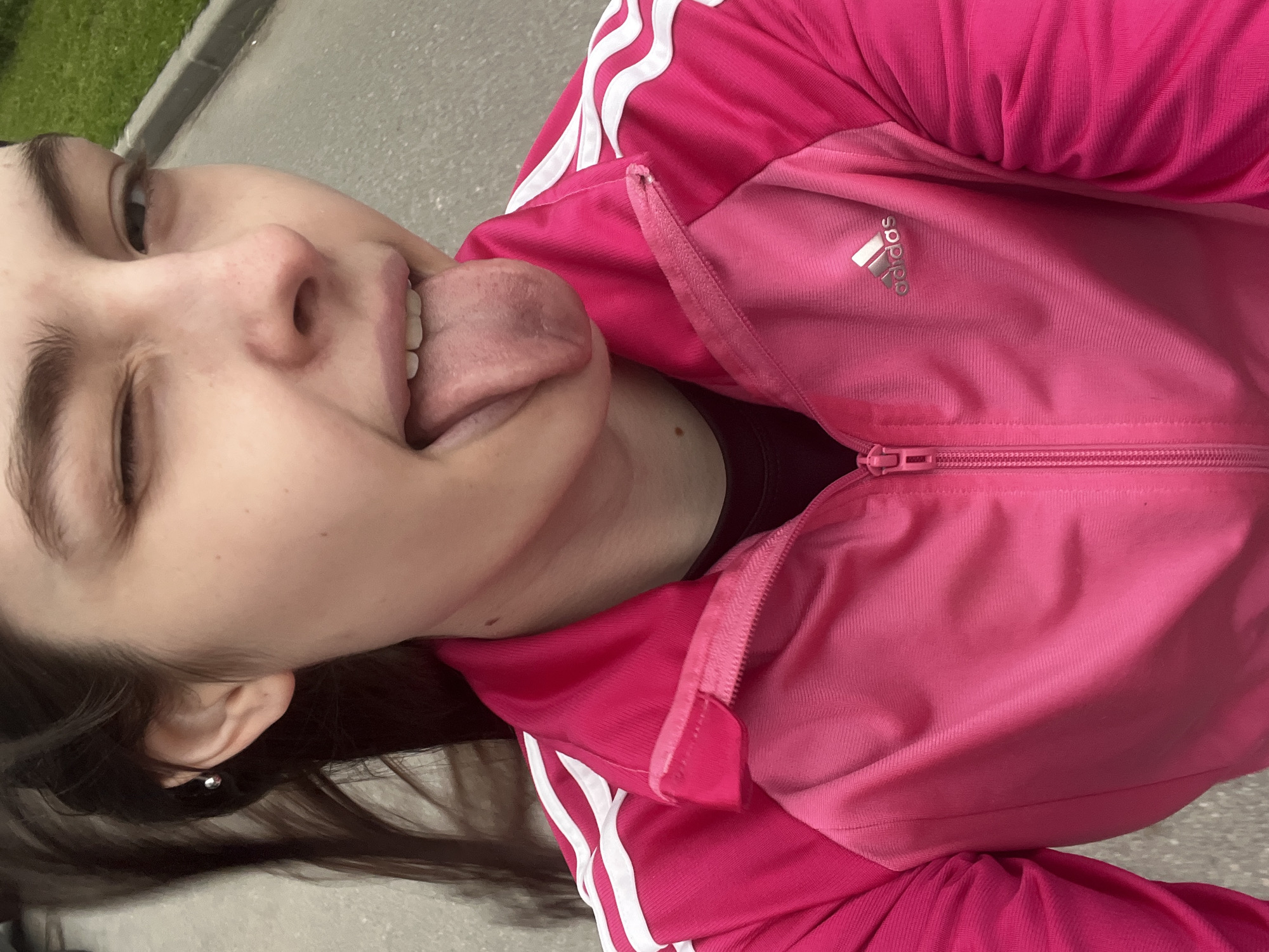 A young woman wearing a pink Adidas jacket with white stripes is sticking her tongue out and winking. She is outside on a paved surface. The location is potentially Saint Petersburg, Russia.