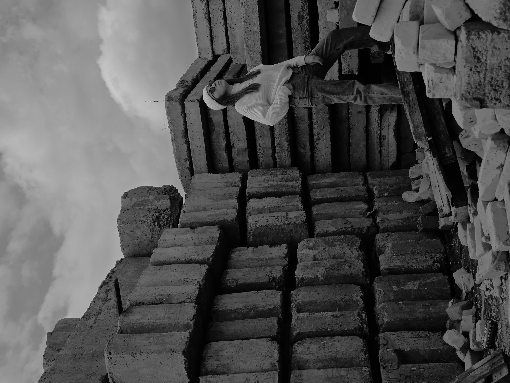 A black and white medium shot photograph depicts a woman in jeans and a sweater laying atop stacked concrete blocks with a cloudy sky in the background. The location is unknown.
The woman is wearing sunglasses and a beanie. She is laying on her side atop the blocks, facing upward.
The blocks are stacked in an irregular fashion, with some of them appearing broken or damaged. There is also debris, including pieces of wood and brick.