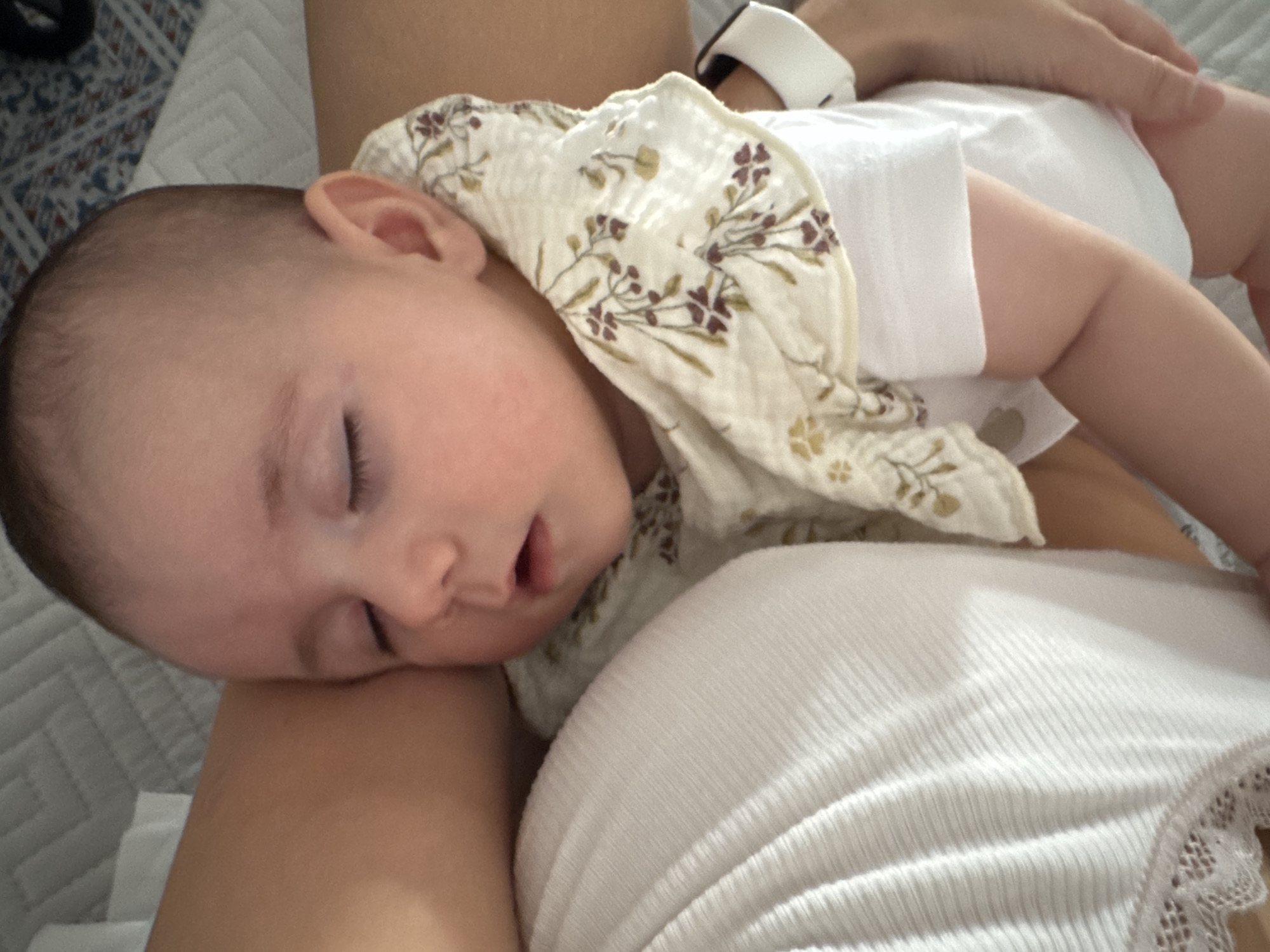 A baby is sleeping on someone's lap. The baby is wearing a white onesie and a bib with a floral pattern. The person's arm is visible on the right side of the image. The background shows a textured surface, possibly a blanket or bedding. The scene is illuminated by natural light, casting soft shadows. The location is not identifiable within the city of Isola delle Femmine, Italy.