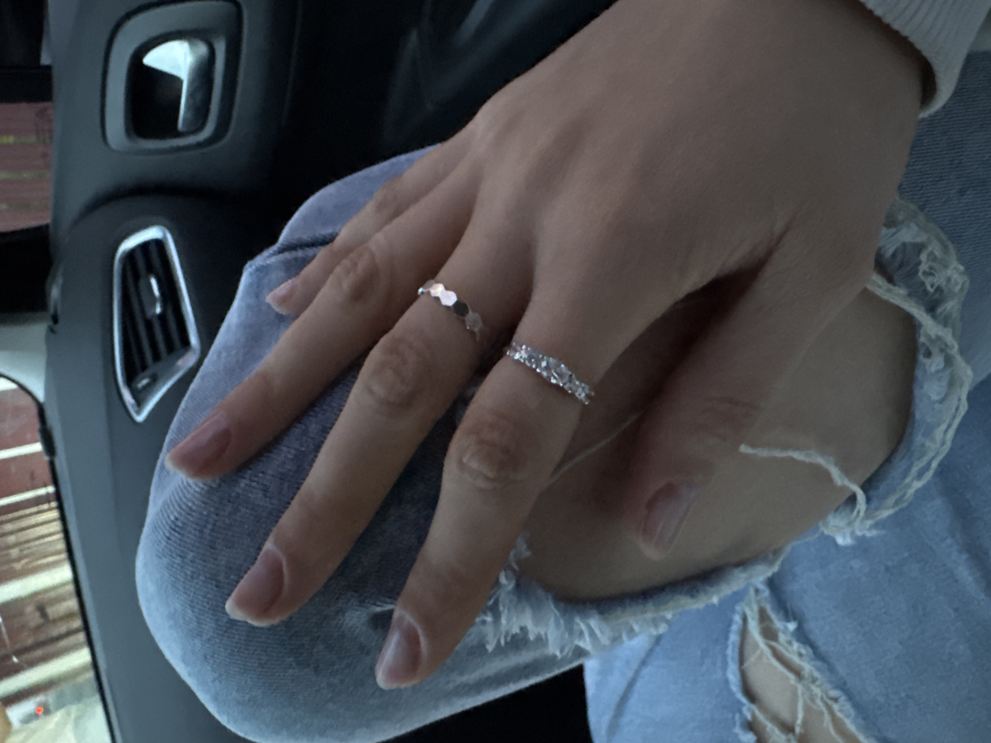 A person's hand, adorned with two rings, rests on a lap clad in ripped denim jeans. The ring on the ring finger appears to be set with small gemstones, while the ring on the middle finger is composed of a series of small metallic discs. The scene appears to be inside a car, evidenced by the dashboard elements visible in the background, including an air vent and a door handle. The location in the city of Mayak, Russia is not possible to ascertain from the image alone.
