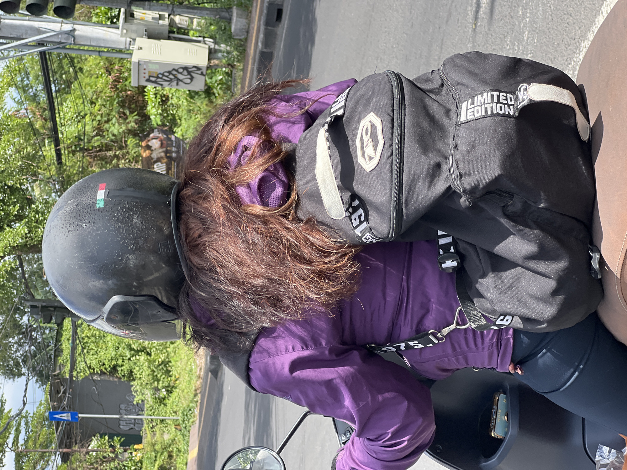 A person wearing a purple jacket and a black helmet rides a scooter. They have a black backpack with "LIMITED EDITION" written on it strapped to their back. The scene appears to be a street in Denpasar, Indonesia, with lush green trees and a traffic light visible in the background. The weather seems to be sunny.