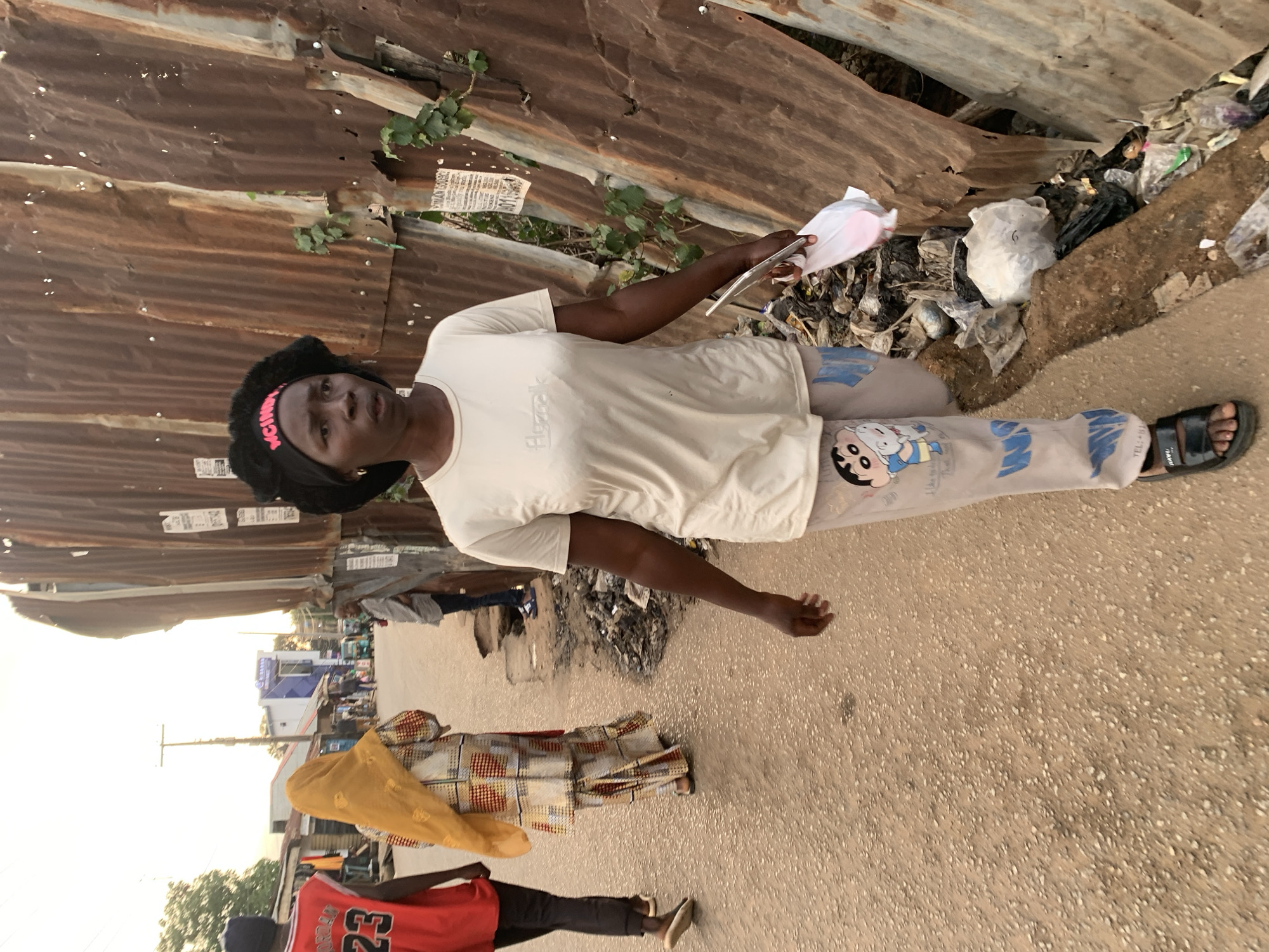 A woman stands on a street in Gura Top, Nigeria, holding papers. She wears a white shirt, gray pants with a cartoon character, and black sandals. A pink headband adorns her head. The backdrop consists of rusted corrugated metal. Litter is scattered nearby. Several people are visible further down the street.