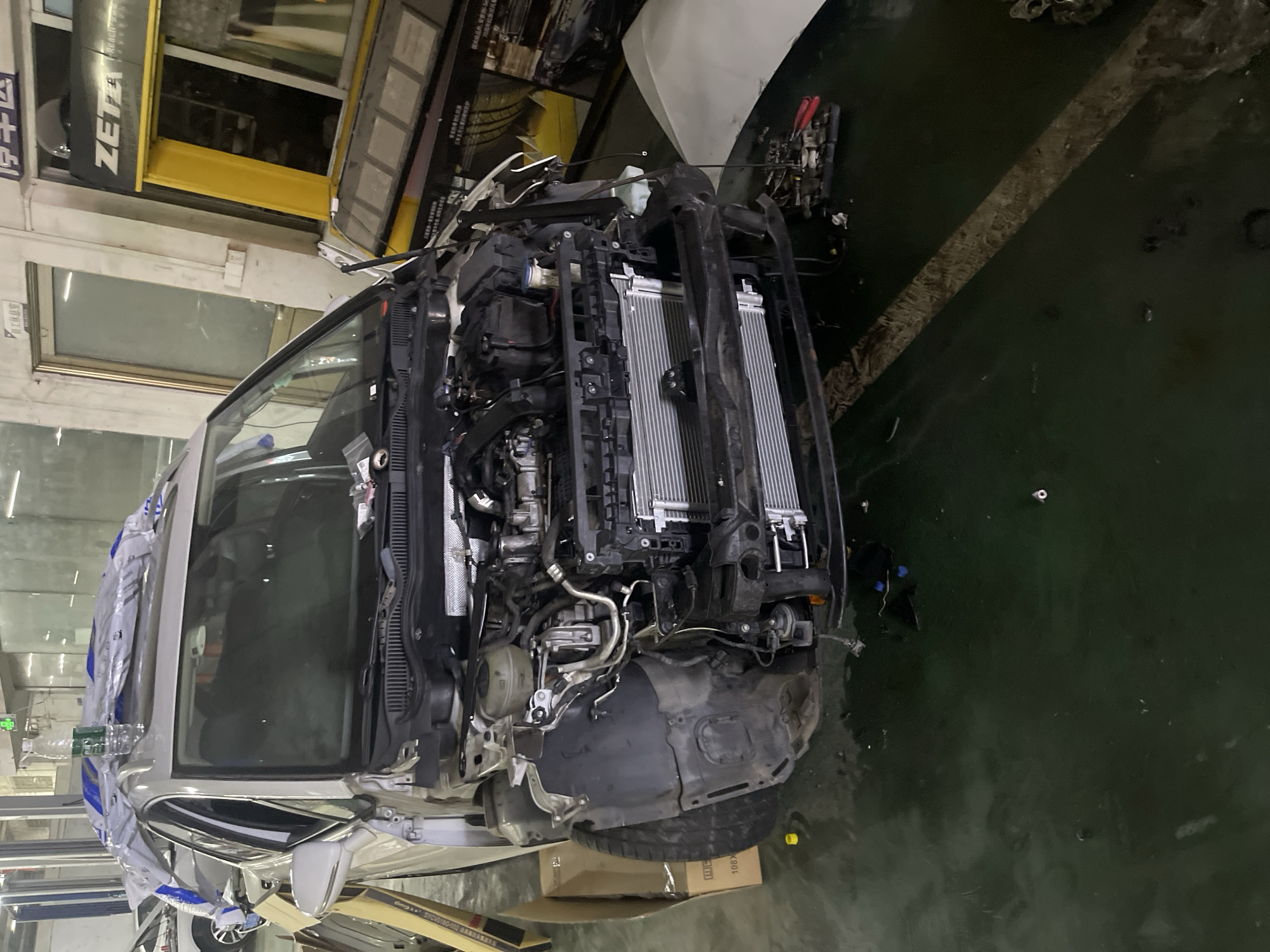 The image shows a white car with its hood open, parked inside a garage or repair shop. The car's front bumper and some of its internal components are disassembled. Several parts are lying on the ground around the vehicle. The garage appears to be industrial, with concrete floors and exposed walls. In the background, there are shelves, machinery, and signage, including the "ZETA" logo. The overall scene suggests that the car is undergoing repairs. The location is identified as Panyu District, China.