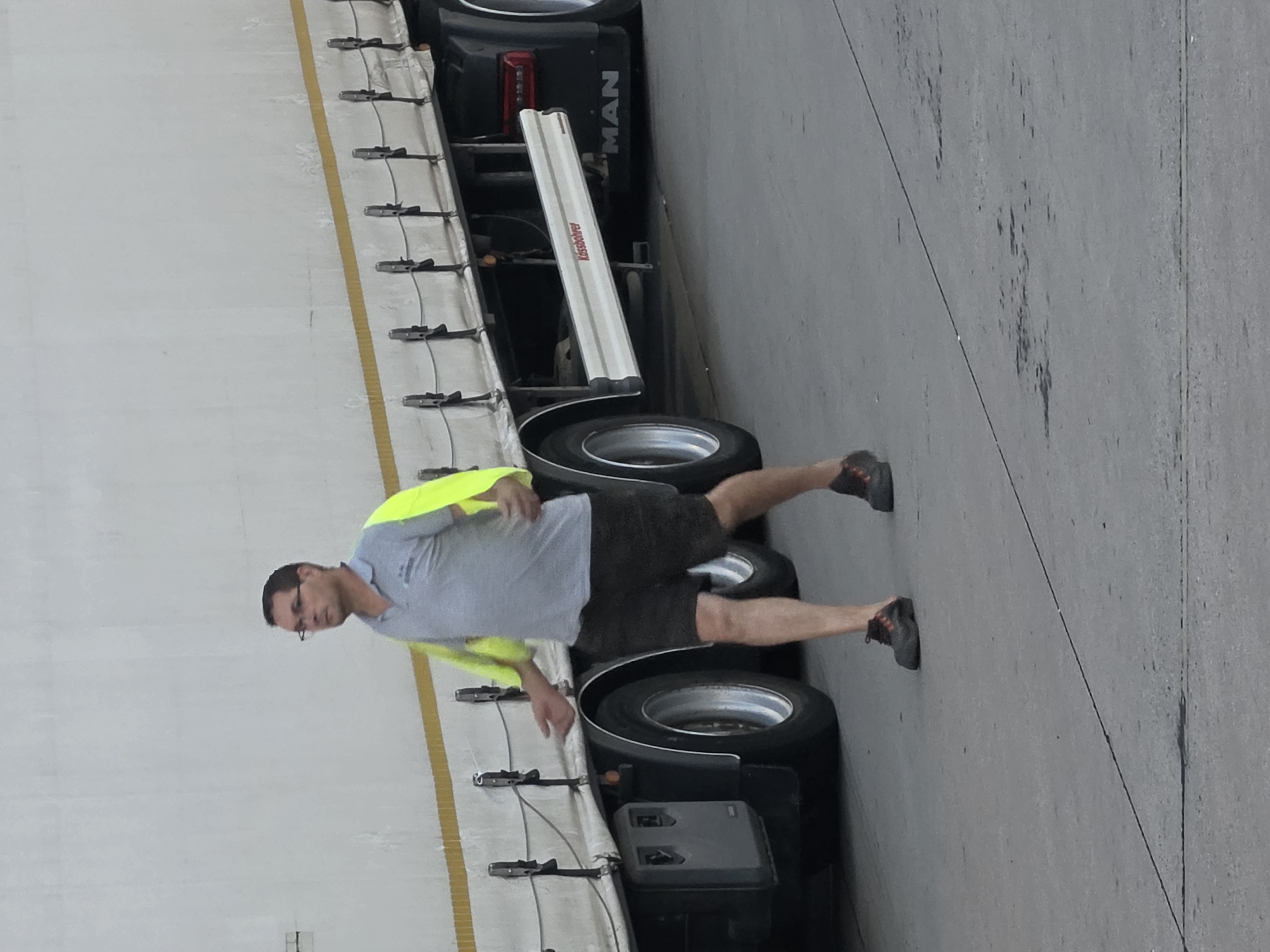 A man wearing glasses, a grey polo shirt with a high-visibility yellow vest over it, and black shorts walks on a paved surface next to a large white truck. The truck has a yellow stripe along its side and visible wheels. The man's right hand is extended outwards, and his left hand is slightly raised. The ground appears to be concrete or asphalt, with some dark stains visible. The scene appears to be in an industrial area or a port, possibly in Frankfurt, Germany, given the presence of the "MAN" logo on the truck.