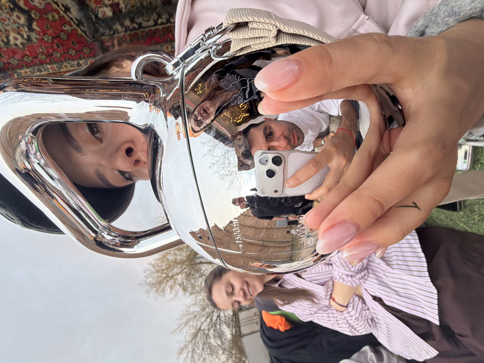 A selfie reflecting in a polished metallic object is held by a person with manicured nails and a small tattoo on their finger. The reflection features a man holding a smartphone, capturing the selfie, and a woman, possibly the person holding the object. The background reflection shows trees, a brick structure marked "1500ml", and other people. In the foreground, another person smiles into the camera. The location appears to be outdoors, possibly Krasnoyarsk, Russia, given the Cyrillic text on the brick structure.