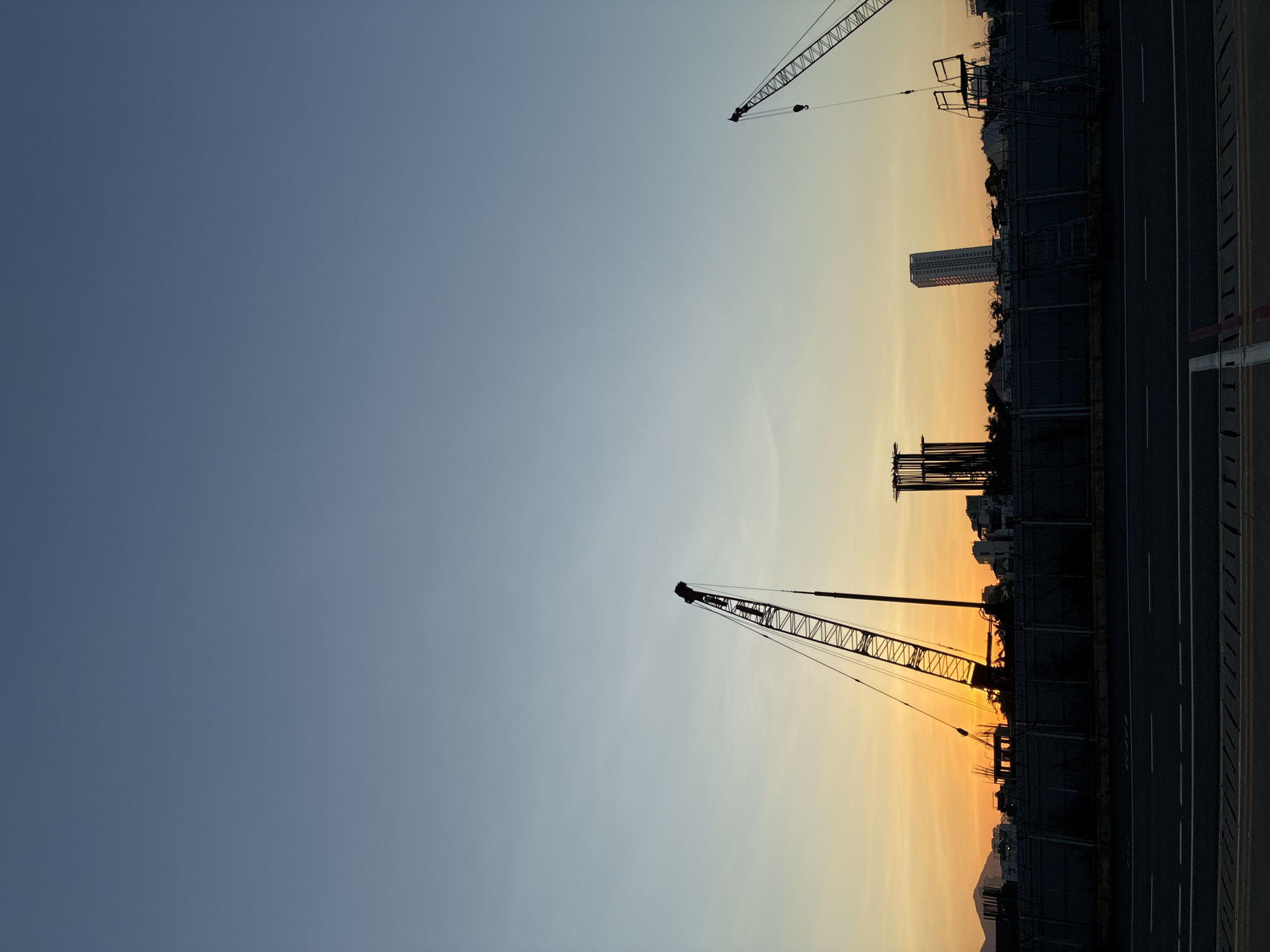 The image depicts a construction site in Da Nang, Vietnam, during what appears to be dawn or dusk. The sky is a gradient of pale blue to a warm orange glow near the horizon, indicating the sun is either rising or setting. Silhouetted against the sky are several large construction cranes. One crane, prominent on the left, is angled upwards, its lattice structure clearly defined. Another crane is visible on the right, with its hook hanging down. In the background, various buildings are silhouetted, including what looks like an unfinished structure with exposed rebar and a tall, slender skyscraper. A dark fence runs across the foreground, separating the construction site from a road. The road itself is asphalt with white lane markings visible. The overall scene is one of urban development and progress set against the natural beauty of the sky.