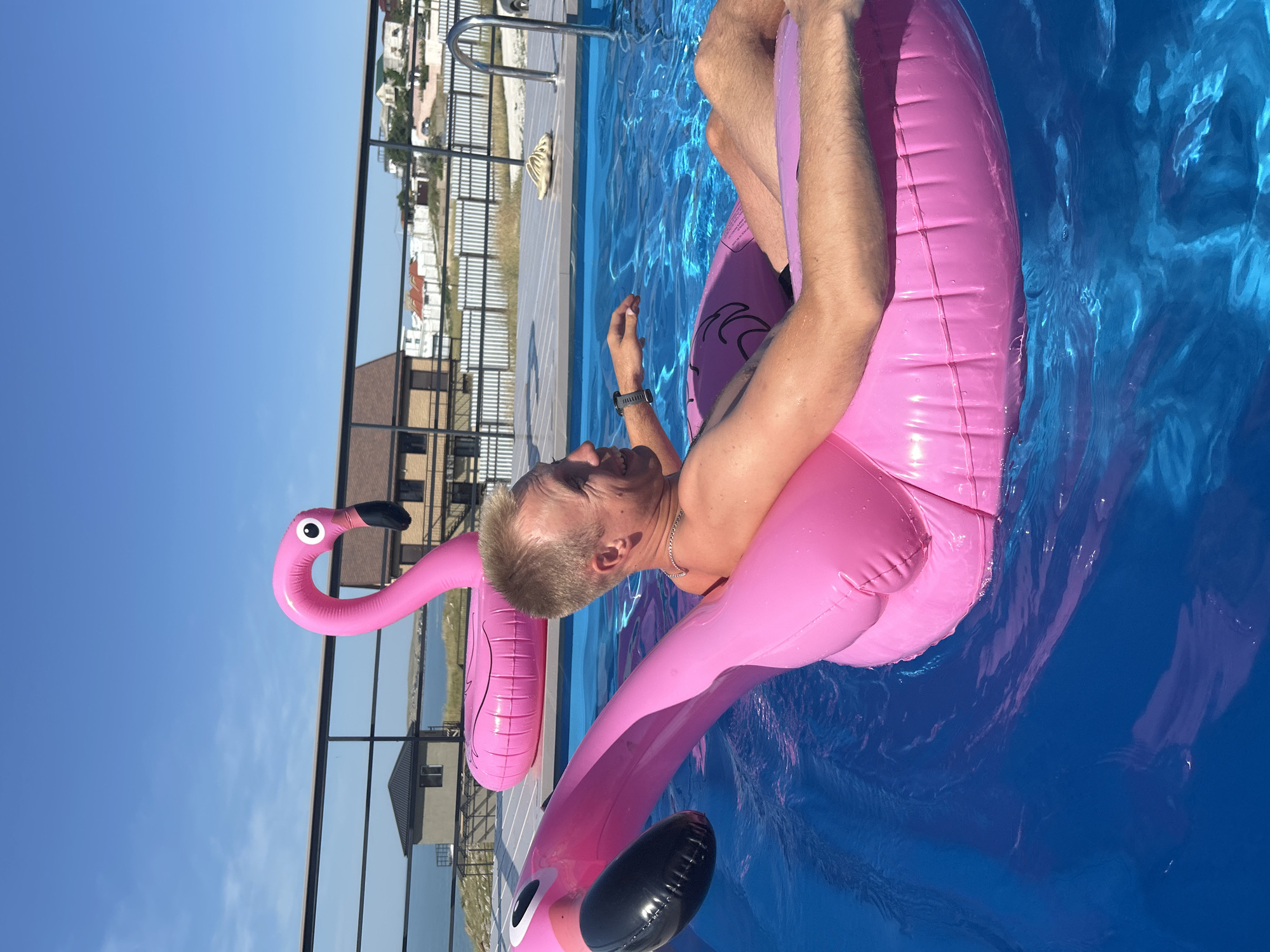 In this medium shot, a man is relaxing in a bright blue swimming pool, floating on a large, inflatable pink flamingo. The man is leaning back on the flamingo, his head resting on its body, and his arms are spread out. He is wearing a silver chain around his neck and a watch on his left wrist. His expression appears content and relaxed.
The flamingo is a prominent object, its neck curved upwards and its body forming a comfortable float. The water is clear and reflects the bright sunlight, creating a shimmering effect. In the background, a fence and some buildings are visible, suggesting an outdoor setting. The sky is a clear, vibrant blue, indicating a sunny day. The overall impression is one of leisure and enjoyment on a warm day. The location context provided suggests this scene is taking place in Mysivske rural settlement, Ukraine.