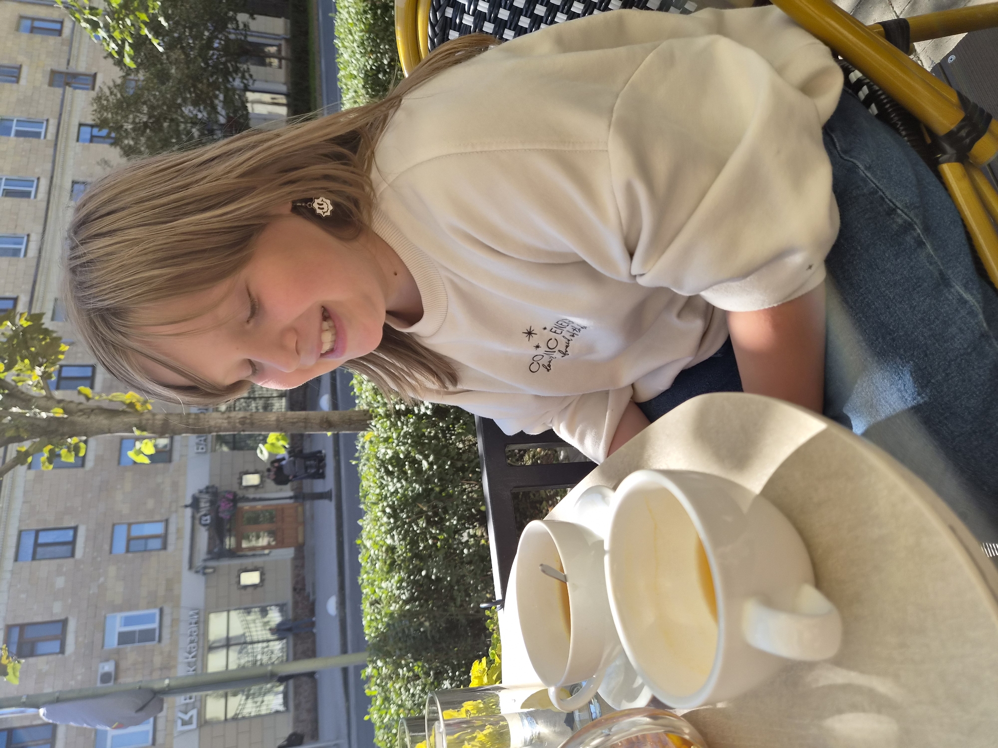 A young girl with blonde hair is sitting at a table outside a cafe in Kazan, Russia. She is wearing a cream-colored sweatshirt and blue jeans. There are two white cups on the table, one with tea and a spoon, and another empty cup. A glass of water with yellow flowers is also on the table. In the background, there is a building with shops and windows, and a street with a sidewalk and trees. The girl is smiling and looking down at the table. The overall scene suggests a casual outdoor dining experience in an urban setting.
