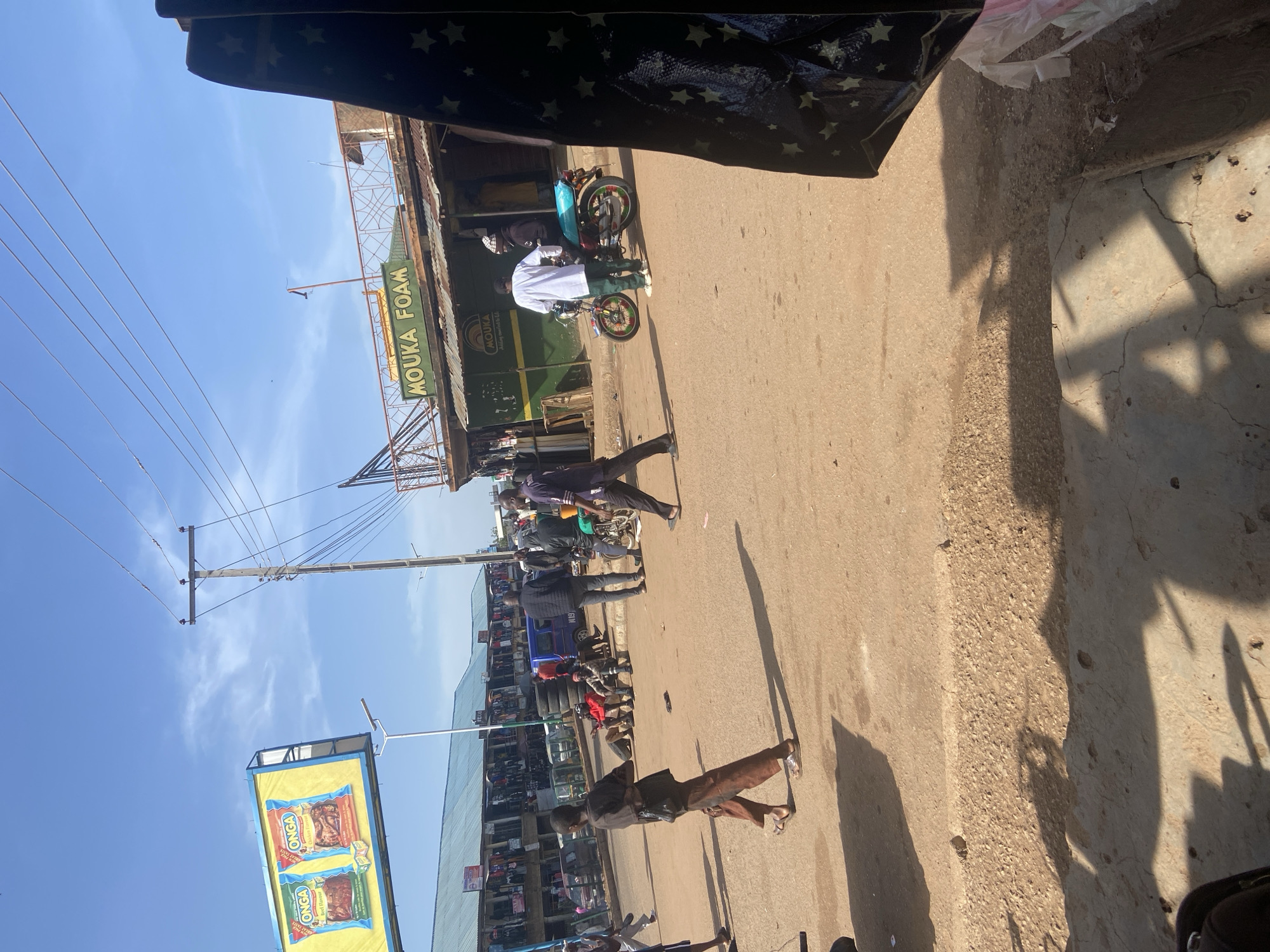 This media file depicts an active street scene in Bukuru, Nigeria, under bright daylight. The image is composed as if viewed from beneath an awning or overhang, showing a paved road with adjacent buildings and numerous individuals.
**Subjects:**
Several people are visible, engaged in various activities. In the midground, a person in a white long-sleeved shirt and dark green trousers is standing next to a red and green motorcycle. Another individual in darker clothing stands further down the street near other parked motorcycles. Towards the foreground, a person in a dark top and brown trousers is walking on the pavement, casting a long shadow. In the background, more people are visible near shops and stalls.
**Activity or event taking place:**
The scene captures daily life on a bustling commercial street, likely a market area. People are walking, standing, and potentially waiting or shopping. Motorcycles, a common mode of transport, are present, suggesting transit and commerce.
**Notable details:**
The time of day appears to be either mid-morning or late afternoon, indicated by the clear blue sky and prominent, elongated shadows cast by people and structures. The weather is sunny and clear. On the left side of the street, utility poles with numerous power lines run across the frame. Buildings lining the street house various businesses. One shop prominently features a green sign with "MOUKA FOAM" written in white capital letters. Below it, part of the name "AOUKA" is also visible, likely related to the brand. Further down the street, a yellow billboard displays two "ONGA" brand seasoning packets, one labeled "Jollof Flavour" and the other "Stew Flavour."
**Visible text:**
The most prominent visible texts are "MOUKA FOAM" on a shop sign and "ONGA" on a billboard, along with the flavor descriptions "Jollof Flavour" and "Stew Flavour." Part of "AOUKA" is also seen below "MOUKA FOAM."