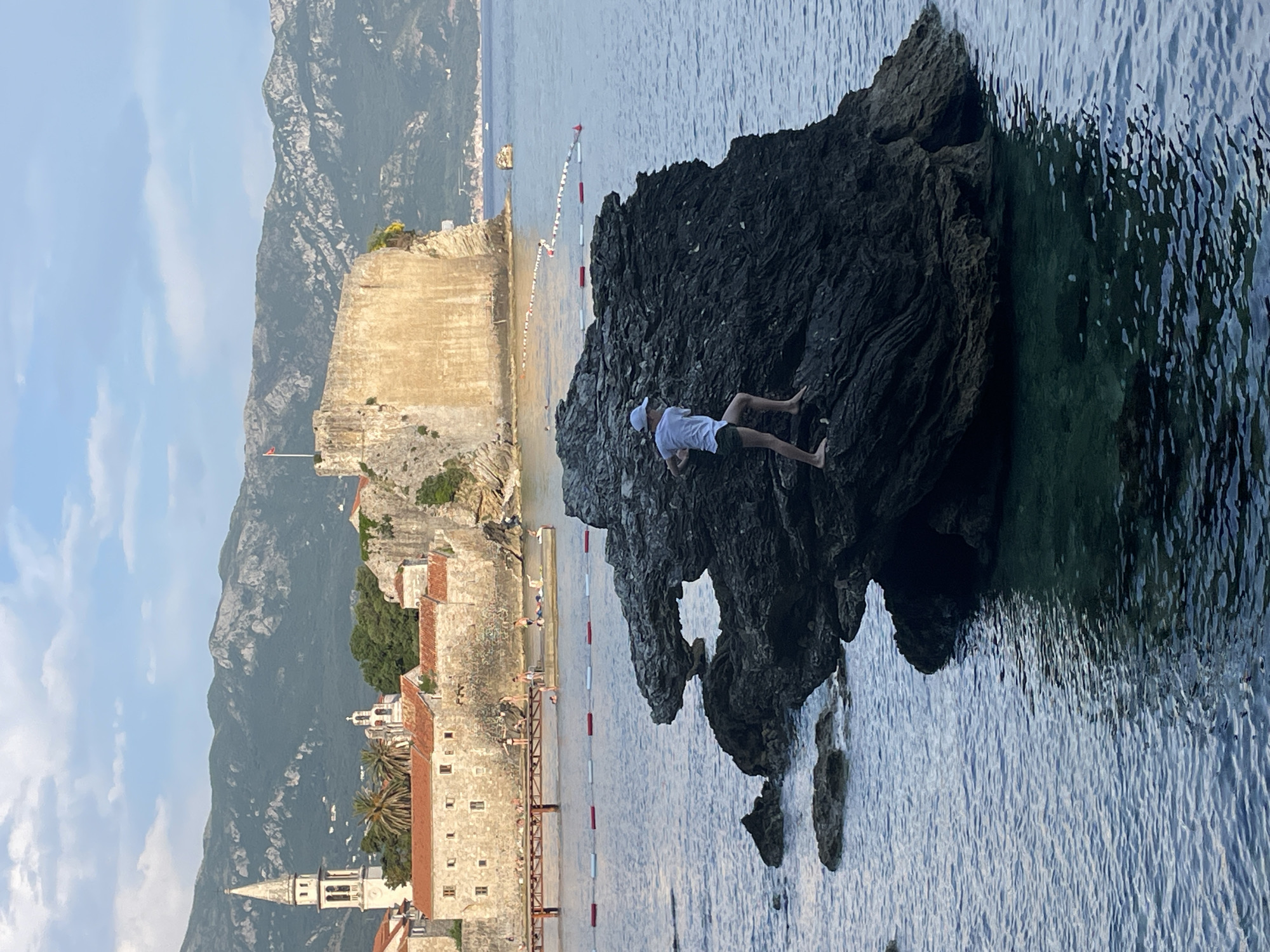 The image depicts a person lying on a large rock formation in the sea near Budva, Montenegro. The person is wearing white clothing and a hat, positioned on the rock with their limbs extended. The rock is located near the coastline, with clear, blue-green water surrounding it. In the background is the historic city of Budva, featuring stone buildings, a church tower, and a medieval citadel along the coast. Mountains are visible in the distance under a blue sky. The scene appears to be taken during daylight with good lighting.