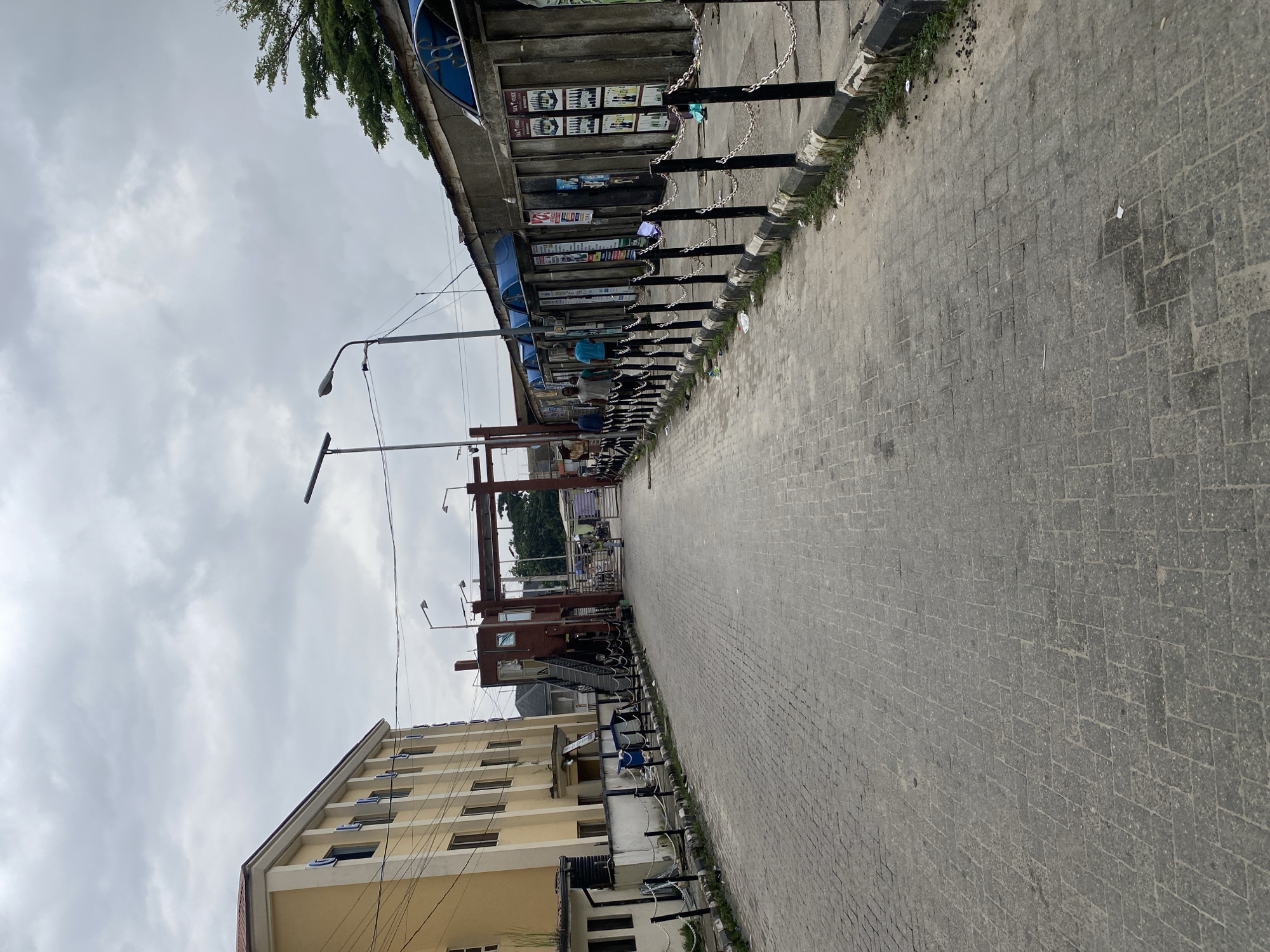 The image shows a paved street with buildings on both sides. On the left is a tall, multi-story yellow building with many windows. On the right, there is a row of small shops with blue awnings. A chain-link fence with black posts separates the shops from the street. Several people are walking down the street, and some are standing near the shops. In the background, there is an archway that appears to be an entrance to another area. The sky is overcast with grey clouds.