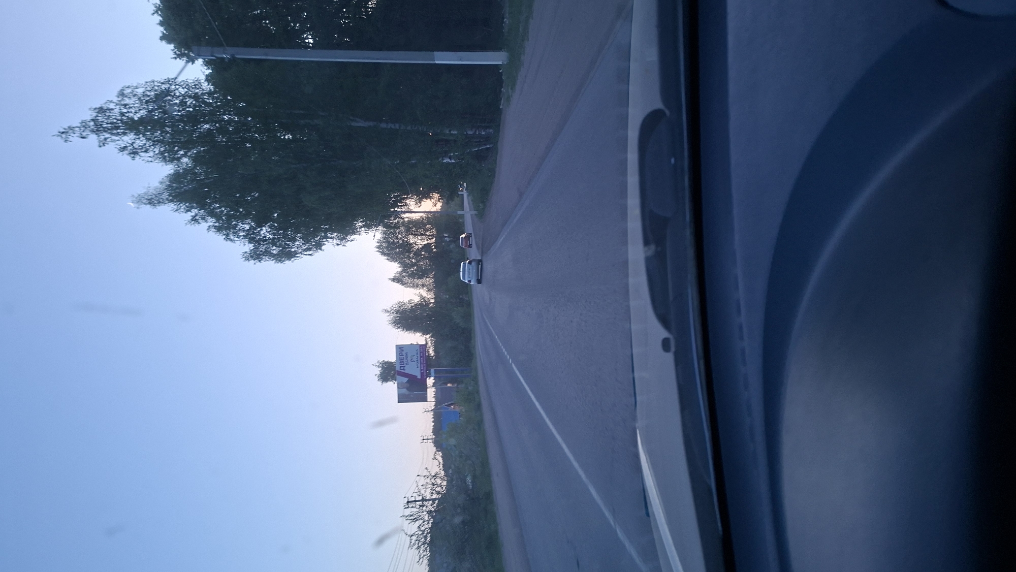 The image depicts a view from inside a vehicle driving along a road. Two cars are visible ahead on the road. There's a billboard on the left side of the road. Based on the Cyrillic lettering on the billboard, the location is identified as Chaikovsky, Russia. Trees line the roadside on both sides. The sky is visible in the upper portion of the image, indicating it's either dawn or dusk. The interior of the vehicle is visible on the right side of the image, including part of the dashboard.
