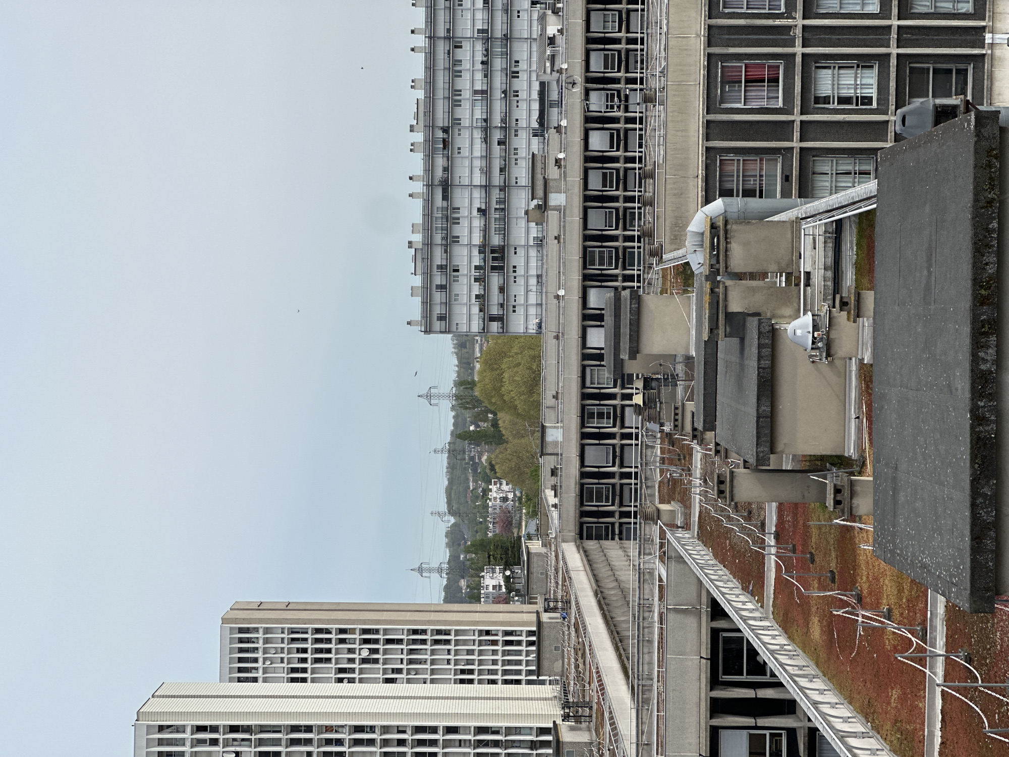 The image depicts an elevated view in Sarcelles, France, likely from a rooftop. Two high-rise apartment buildings dominate the scene, constructed with a modular, repetitive window pattern. The building to the left is lighter in color, while the other shows a darker, possibly older, facade. Various rooftop structures are visible, including ventilation systems, access walkways, and a section of vegetation, possibly a green roof, which is reddish-brown. In the background, power lines traverse the horizon, and a residential area with houses and trees can be discerned. The sky is overcast. No people are visible.