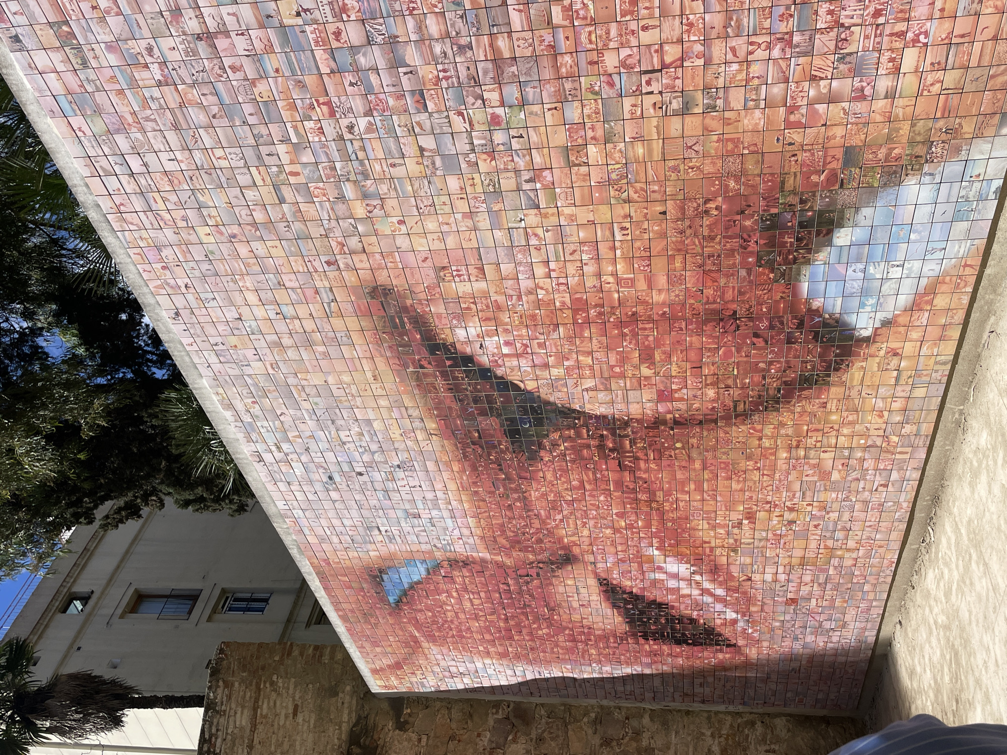 The image shows a large mosaic mural in Barcelona, Spain. The mural is composed of hundreds of small square tiles, each containing a separate image. When viewed together, these tiles create a larger image of a woman's face. The face appears to be in a state of emotional expression. The tiles feature a variety of images in a range of styles. The mosaic is affixed to the underside of a cantilevered structure. The overall structure is made of light-colored concrete. To the left of the mosaic is a tree and a building. The building appears to be a multi-story residential structure with multiple windows and possibly a balcony or other protruding architectural detail. There is also a palm tree. Below the mosaic, the wall is made from old, weathered stone.