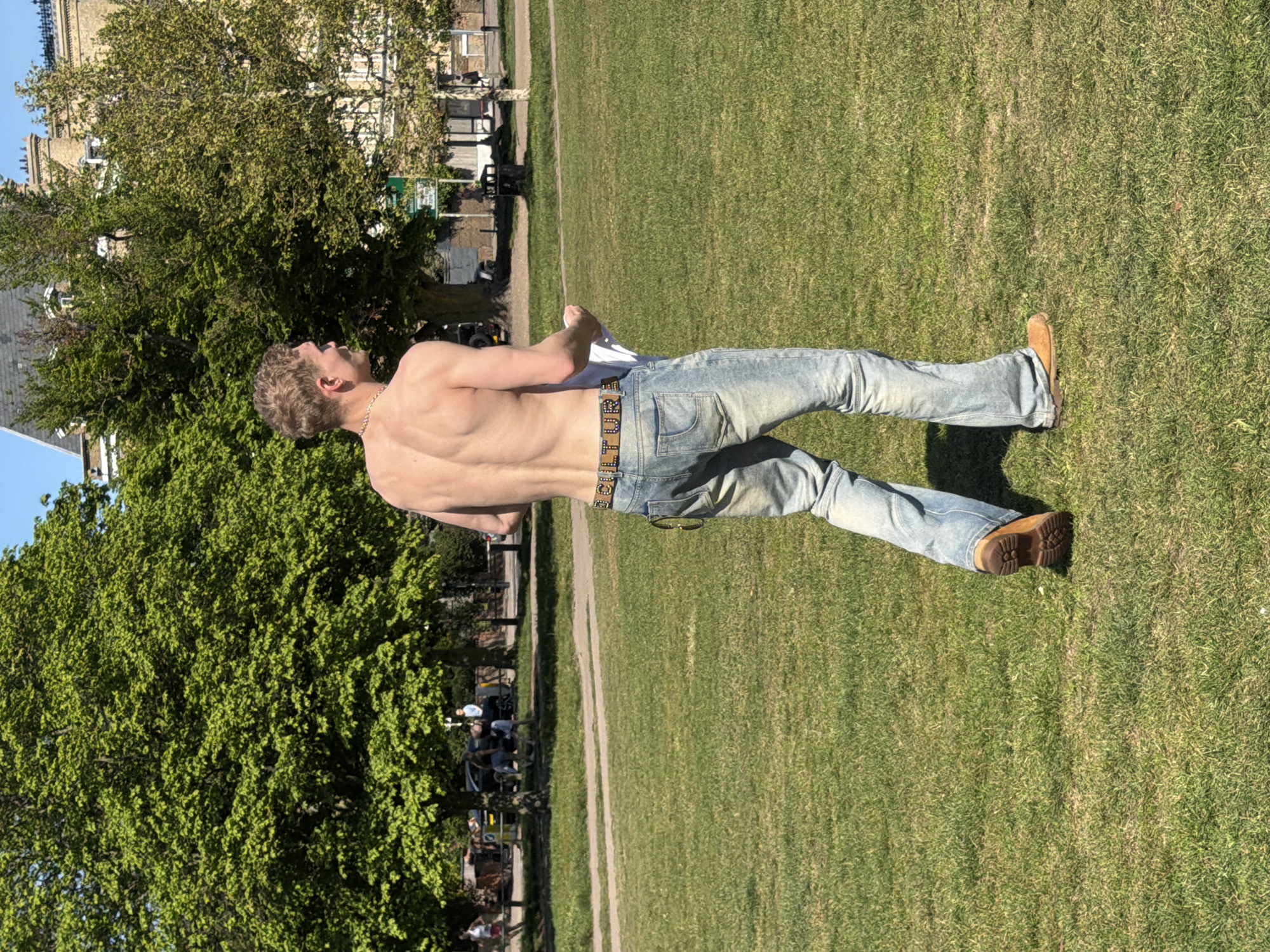 A man with fair hair, wearing blue jeans, a distinctive belt, and brown boots, stands shirtless in a grassy park. He holds a white garment in his left hand. The park is shaded by large trees, and a pathway borders the grass. In the background, there are parked cars and a building. The photo appears to be taken in a public park in London, United Kingdom.