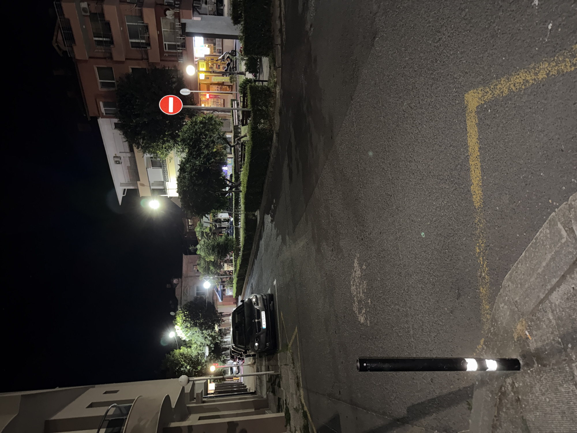 A street scene at night in an urban environment. A dark-colored sedan is parked parallel to the sidewalk on the left side of the image. A black bollard with reflective bands is positioned in the foreground on the left. Yellow painted lines are visible on the asphalt road. In the background, there are buildings, trees, benches, and streetlights. A red and white "no entry" sign is displayed on a pole. A motorcycle is parked near a storefront. The lighting suggests it is nighttime.