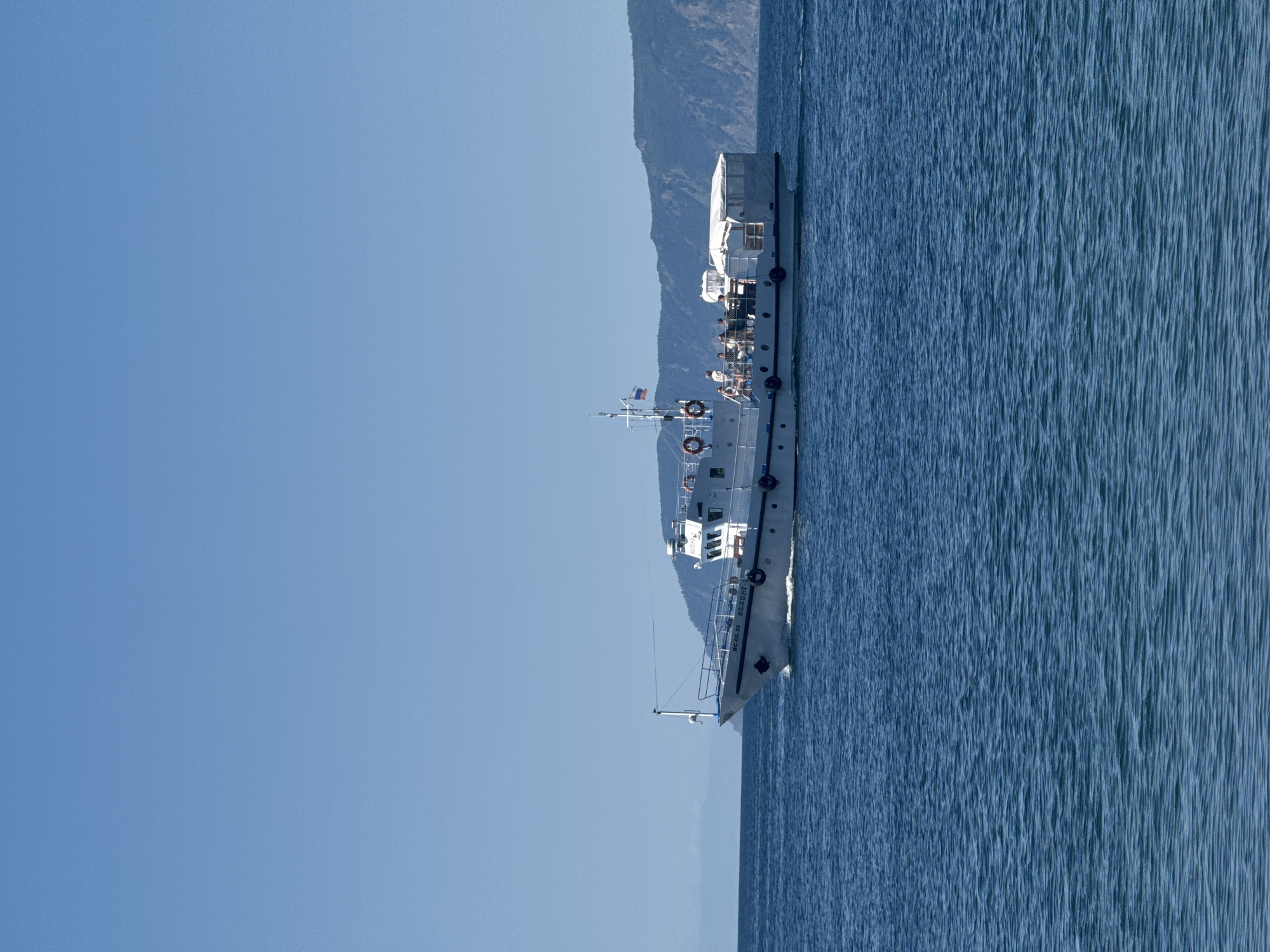 A white tourist boat with blue railings is moving across the deep blue water of Lake Baikal. The boat has several passengers on its upper deck, some of whom are standing and looking out at the scenery. A white canopy covers the rear portion of the deck. The boat is labeled with Cyrillic text and the number "BC-05-30". In the background, a densely vegetated mountain range with a distinct ridgeline rises from the far shore of the lake. The sky above is clear and bright blue. This scene is consistent with the typical landscape and tourist activities observed in Listvyanka, Russia, a settlement on the western shore of Lake Baikal.