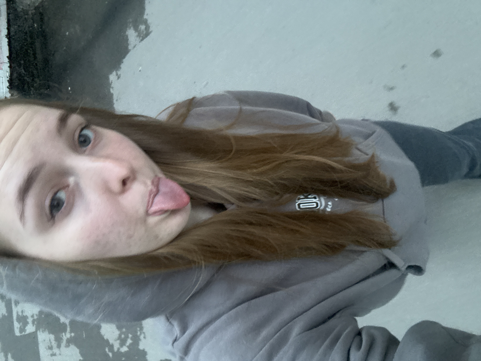 A young woman is depicted in a selfie-style image. She has fair skin, light eyes, and long brown hair. She is wearing a gray hooded sweatshirt. Her tongue is extended, creating a playful expression. The background appears to be a textured surface, potentially concrete. The photo was taken in Perm, Russia.