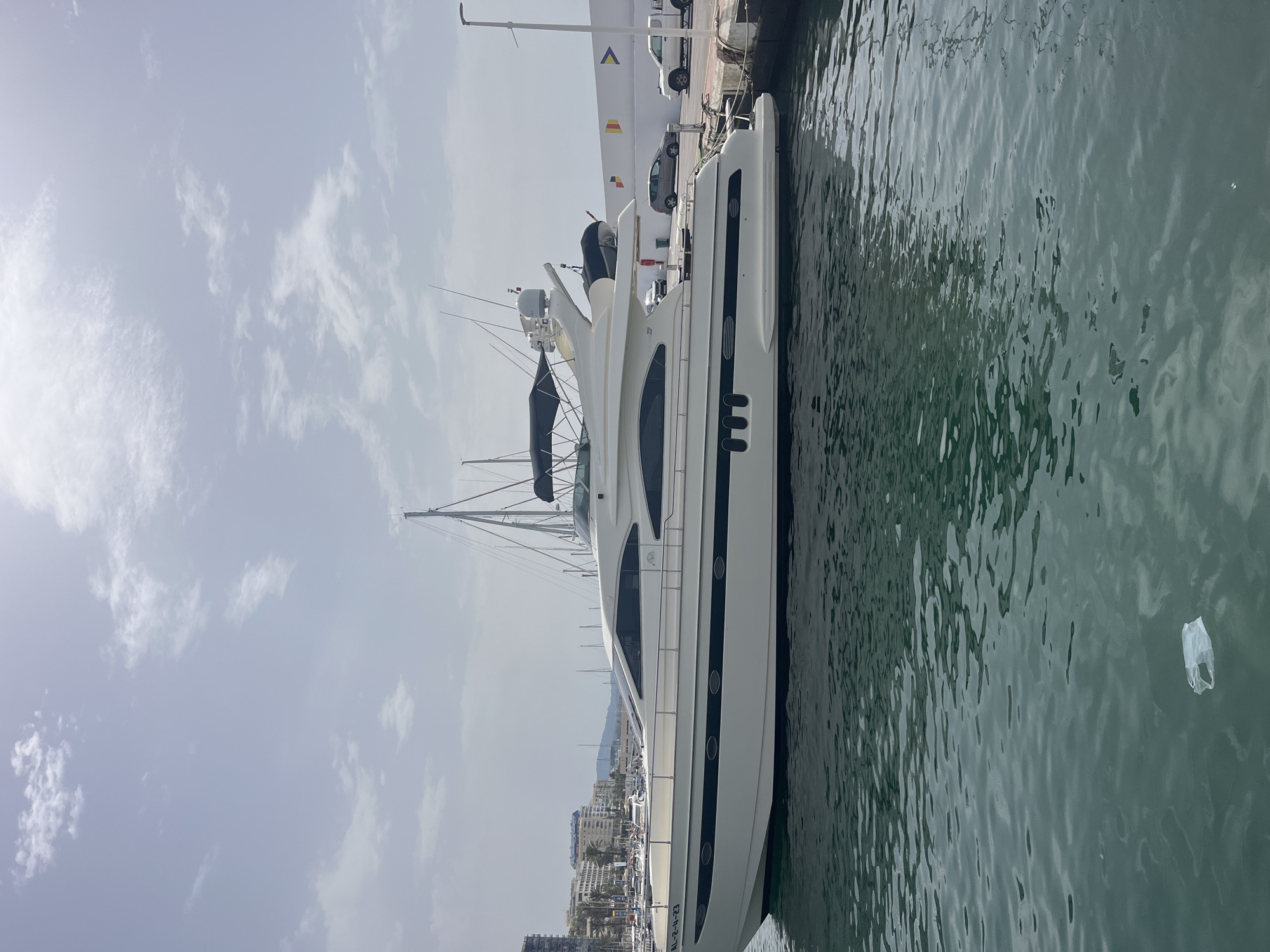 The image depicts a large, white motor yacht docked in a marina in Marbella, Spain. The yacht is the central focus, with its sleek lines and multiple decks visible. The water in the foreground is a deep green, with ripples and a floating plastic bag. In the background, other boats and yachts are moored, along with buildings and palm trees characteristic of a coastal city. A white wall with colored flags and a street lamp borders the marina. Cars are parked along the waterfront. The sky is overcast with scattered clouds. The overall scene suggests a sunny day in a popular Mediterranean tourist destination.