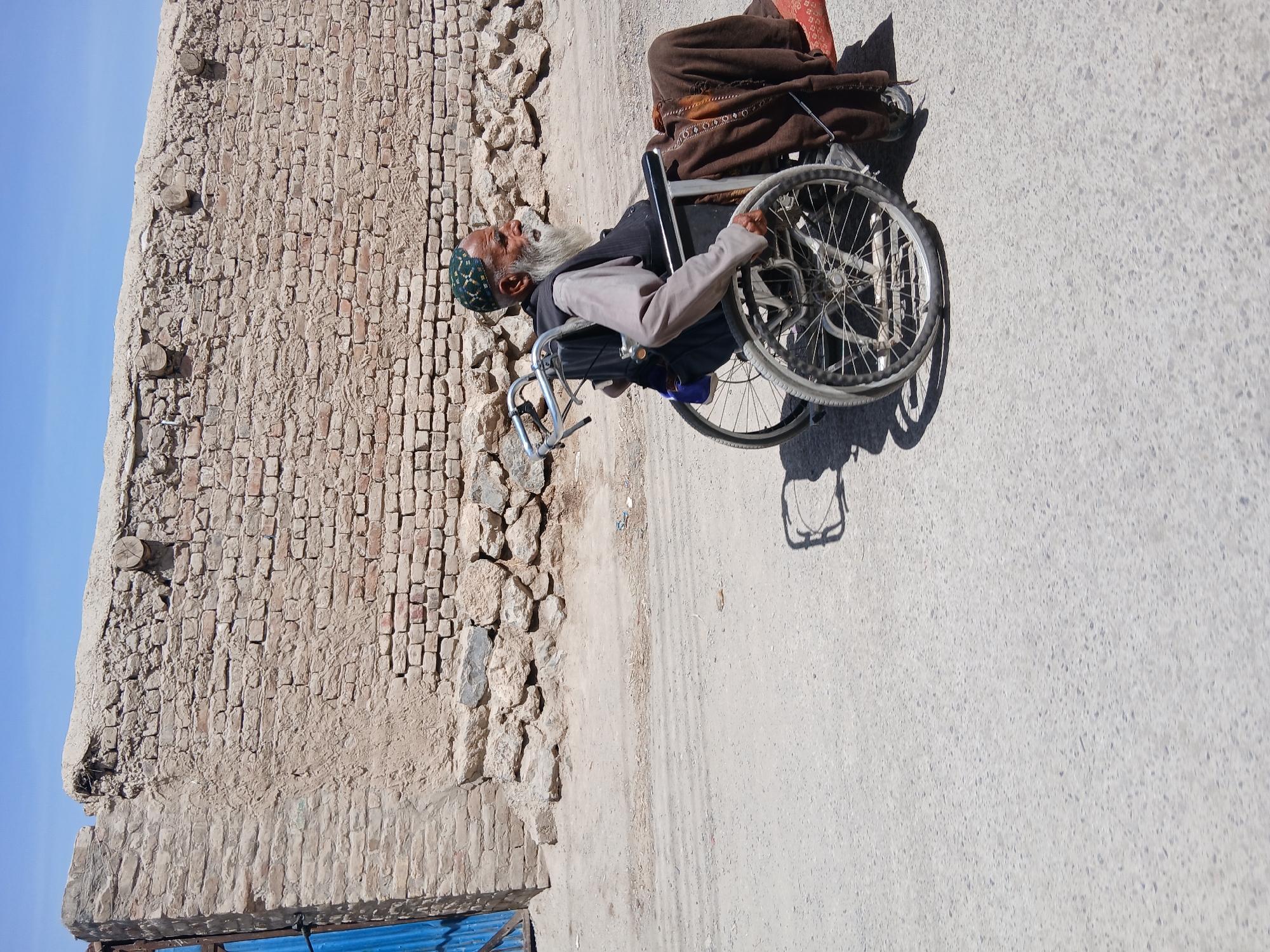 The image features an elderly man with a long white beard, wearing a green skullcap and a grey jacket over a dark garment, seated in a manual wheelchair. His eyes are closed or looking upwards, with a serene expression. A brown blanket or fabric covers his lower body. His right hand rests on the rim of the wheelchair's large wheel.
The scene is outdoors, set in Quetta City Tehsil, Pakistan, under a clear blue sky. To the man's left, a tall, weathered wall constructed primarily of mud bricks and rough-hewn stones rises from the ground. Parts of the wall show signs of crumbling and repair, with a section of blue corrugated metal visible at its base. The ground surface is a mix of light-colored dirt and a paved area. Sharp shadows cast by the wheelchair indicate bright sunshine, suggesting it is mid-day. The man appears to be resting or waiting. No visible text is present in the image.