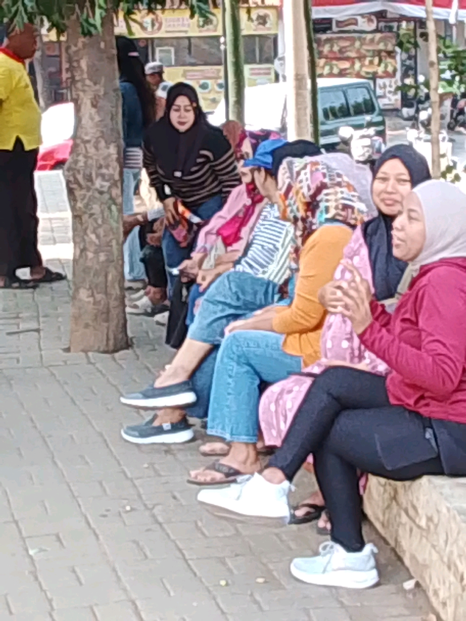 The image depicts a group of people, primarily women, seated and standing outdoors. The setting appears to be a public area with a paved walkway and a tree in the foreground. In the background, there are stalls or shopfronts, suggesting a marketplace or a busy street. The presence of a green van and motorcycles further indicates an urban environment. The people are dressed in casual clothing, with several women wearing headscarves. Some are sitting on a raised concrete structure, while others are standing or sitting on the ground. The overall scene suggests a social gathering or a moment of rest in a public space within Probolinggo, Indonesia.