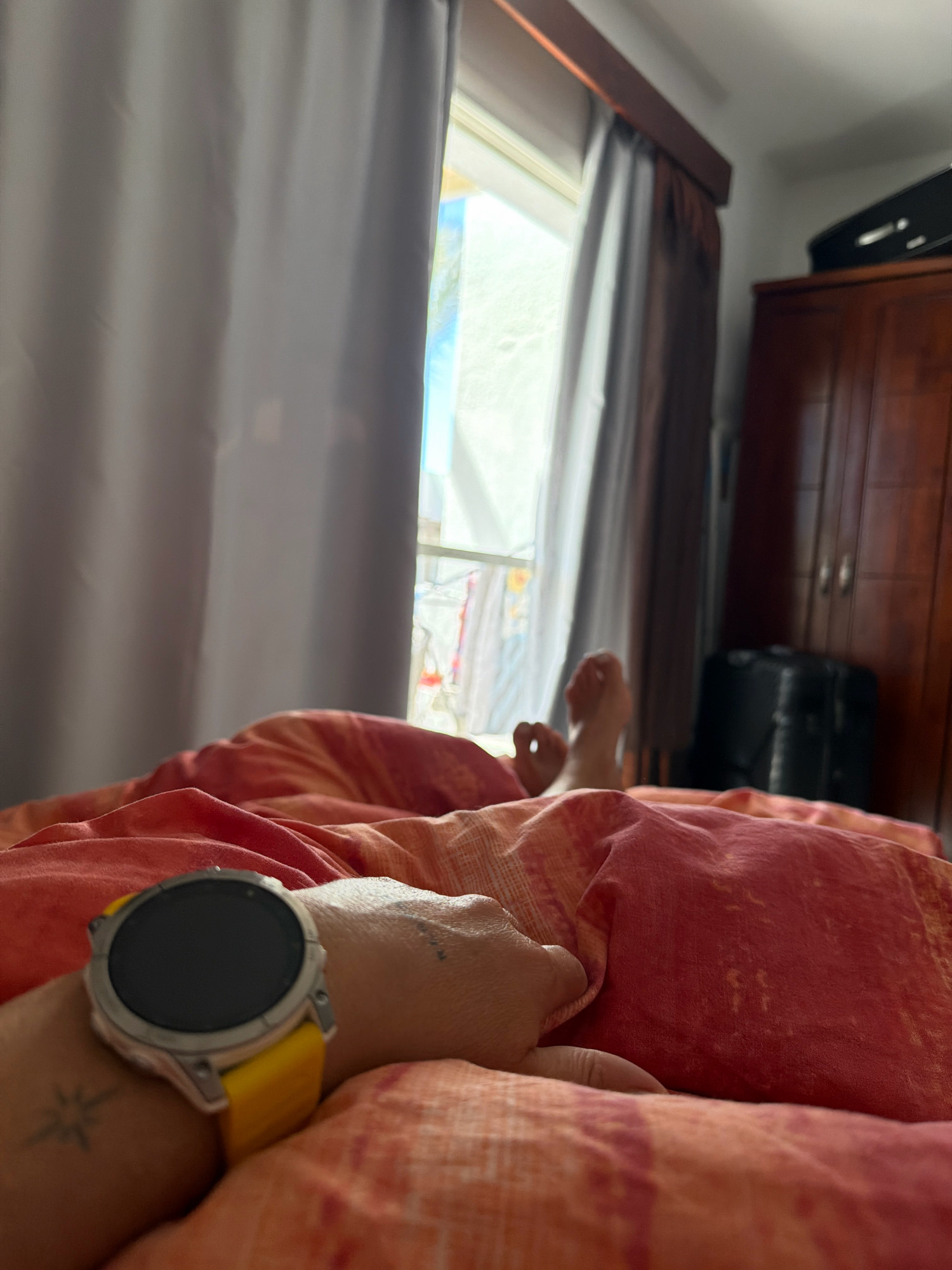 An overhead shot shows a person lying in bed, covered by orange and pink patterned blankets. A hand wearing a silver and yellow smartwatch is resting on the blanket. In the background, curtains are drawn back to reveal a window with a view of palm trees. To the right, a wooden wardrobe and a black suitcase are visible. The setting appears to be a bedroom in Sharm El Sheikh, Egypt.