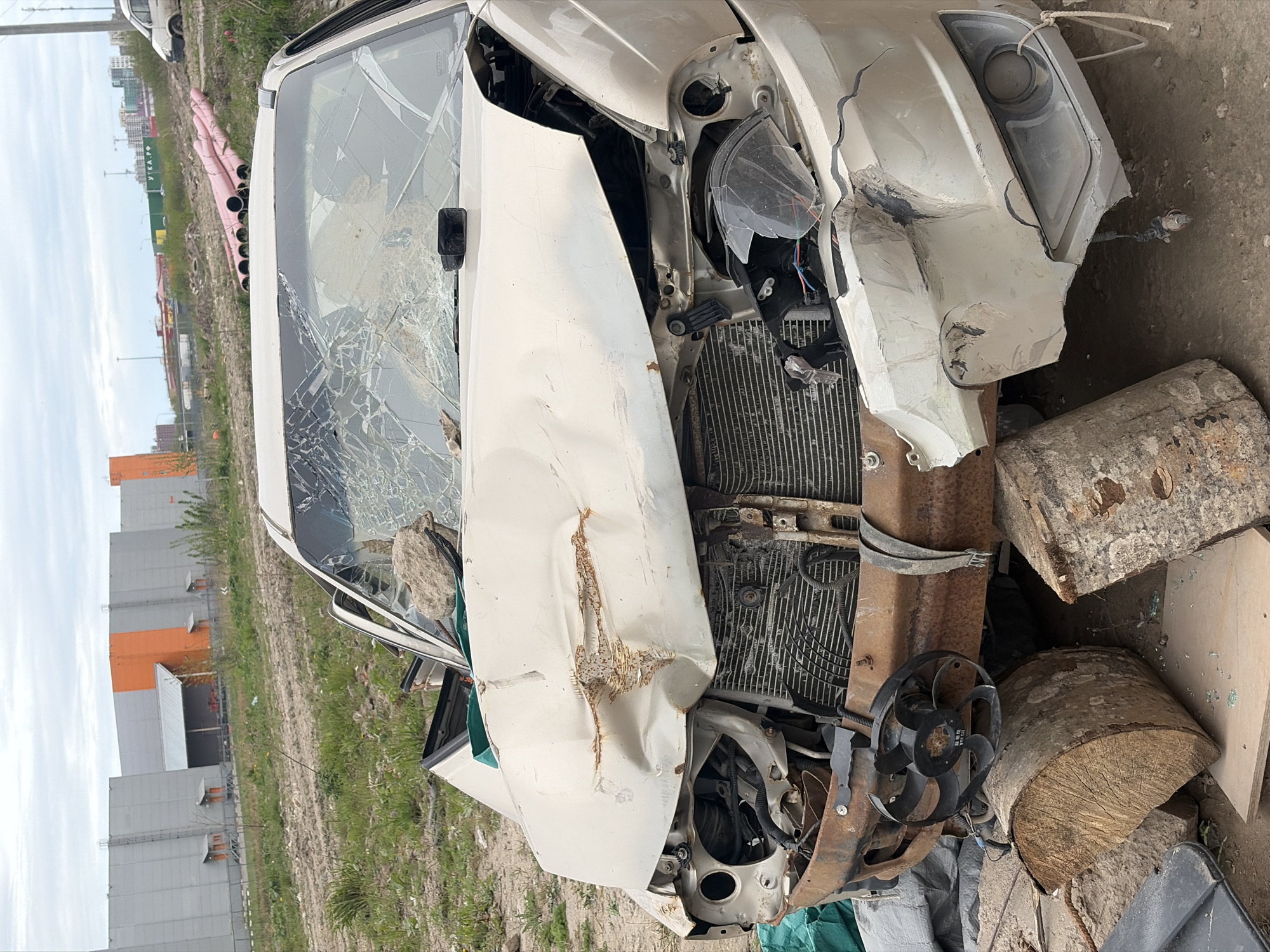 A severely damaged white car is situated in what appears to be a junkyard or abandoned lot. The windshield is shattered, and the front end is crushed, exposing the radiator and engine components. Two cut logs are positioned near the front of the car. In the background, industrial buildings are visible, along with a row of pink pipes. The scene appears to be in an urban or semi-urban area, possibly in Saint Petersburg, Russia. The setting includes bare ground with sparse vegetation. The car is the primary subject, indicating a possible accident or collision. No people are visible.