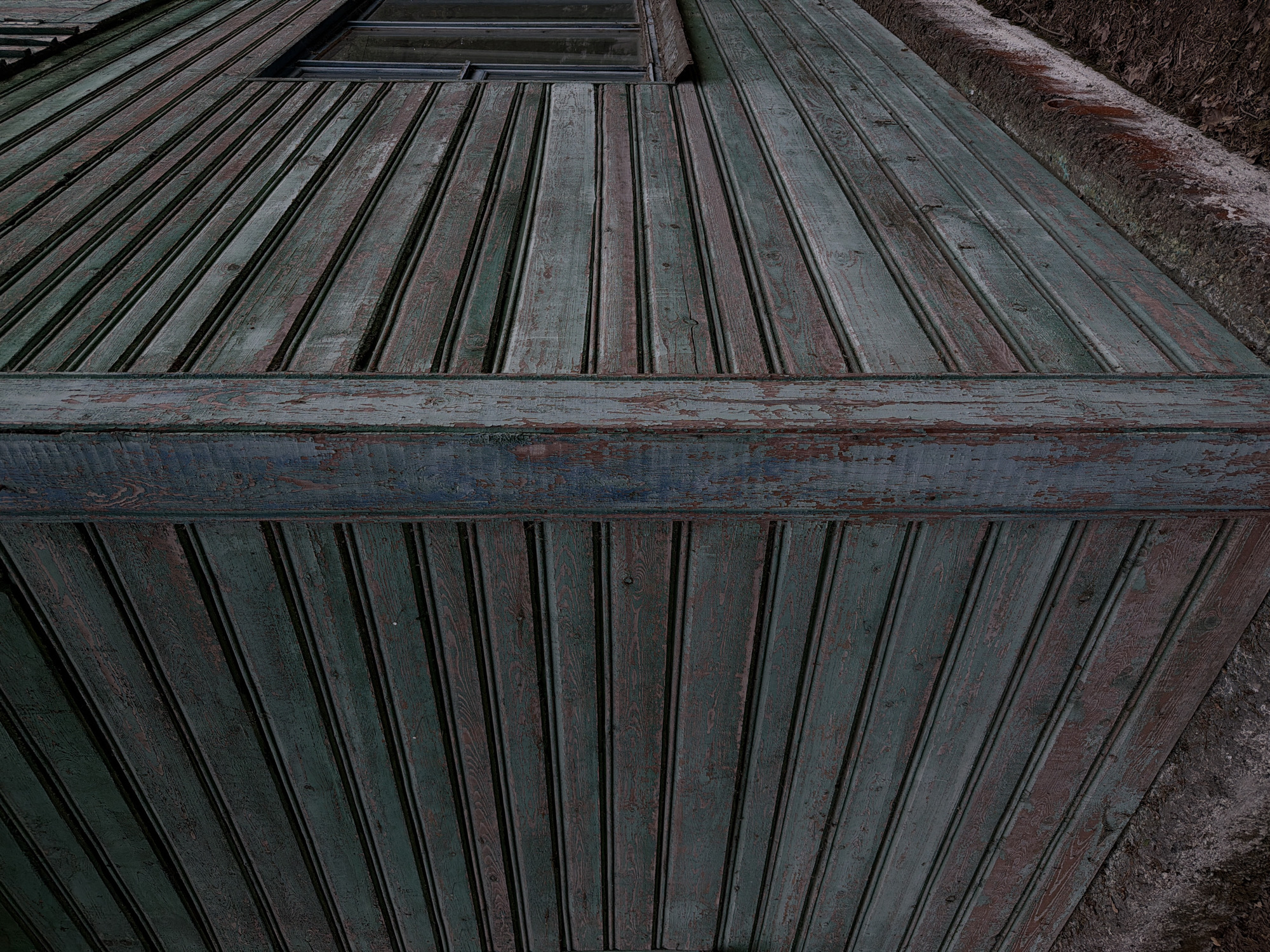 The image shows the weathered exterior of a wooden building, likely a shed or an old house, in the Ракітніцкі сельскі Савет, Belarus. The building's walls are constructed from horizontal wooden planks painted a dark, desaturated green, which is peeling in many areas, revealing reddish-brown underlayers. A vertical wooden beam, also showing signs of wear and peeling paint, forms a corner of the structure. To the right, a small window with a dark frame is visible, set into the wooden siding. The base of the building rests on a rough, concrete-like foundation, which meets the ground covered in dry leaves and debris. The overall impression is one of age, neglect, and the passage of time, with the muted colors and visible decay suggesting an outdoor setting in a rural or semi-rural environment. There are no people or animals present in the image.