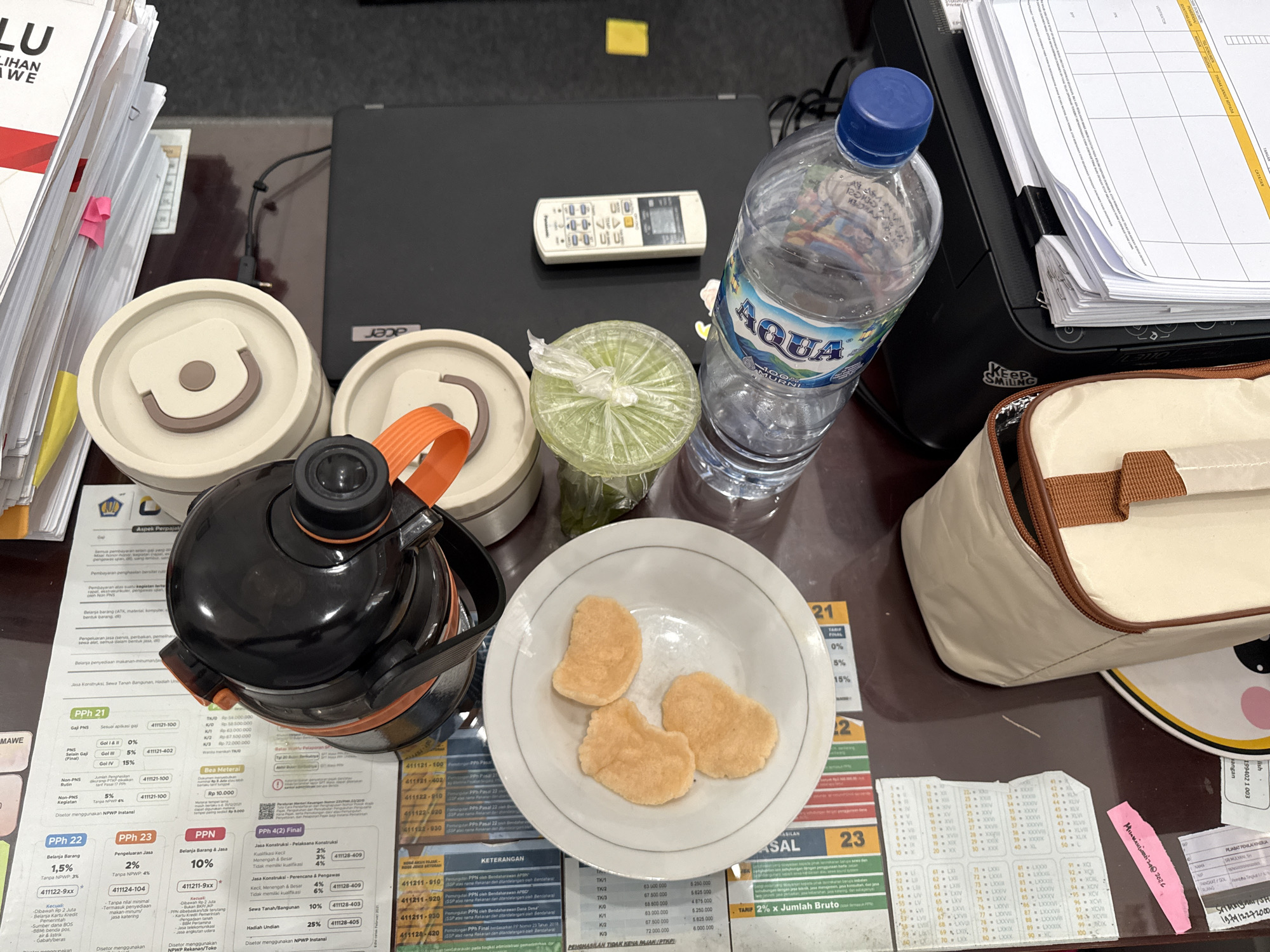 The image depicts a desk or table setting in an indoor environment. Predominant objects include documents (presumably related to finance), a laptop, a remote control, a bottle of Aqua water, food in a bowl, and several containers, including what appears to be a thermos-like food container. A printer is visible at the edge of the frame. The documents are filled with numbers and information, possibly related to taxes (PPN and PPh). The food in the bowl appears to be crackers. A sticker or handwritten note is also present on the desk. There's no indication of the location, but based on the text in the documents and the Aqua bottle's brand, the image is most likely from Indonesia. No people or specific actions are visible. The city of Lhokseumawe, Indonesia is not visibly indicated in the image.