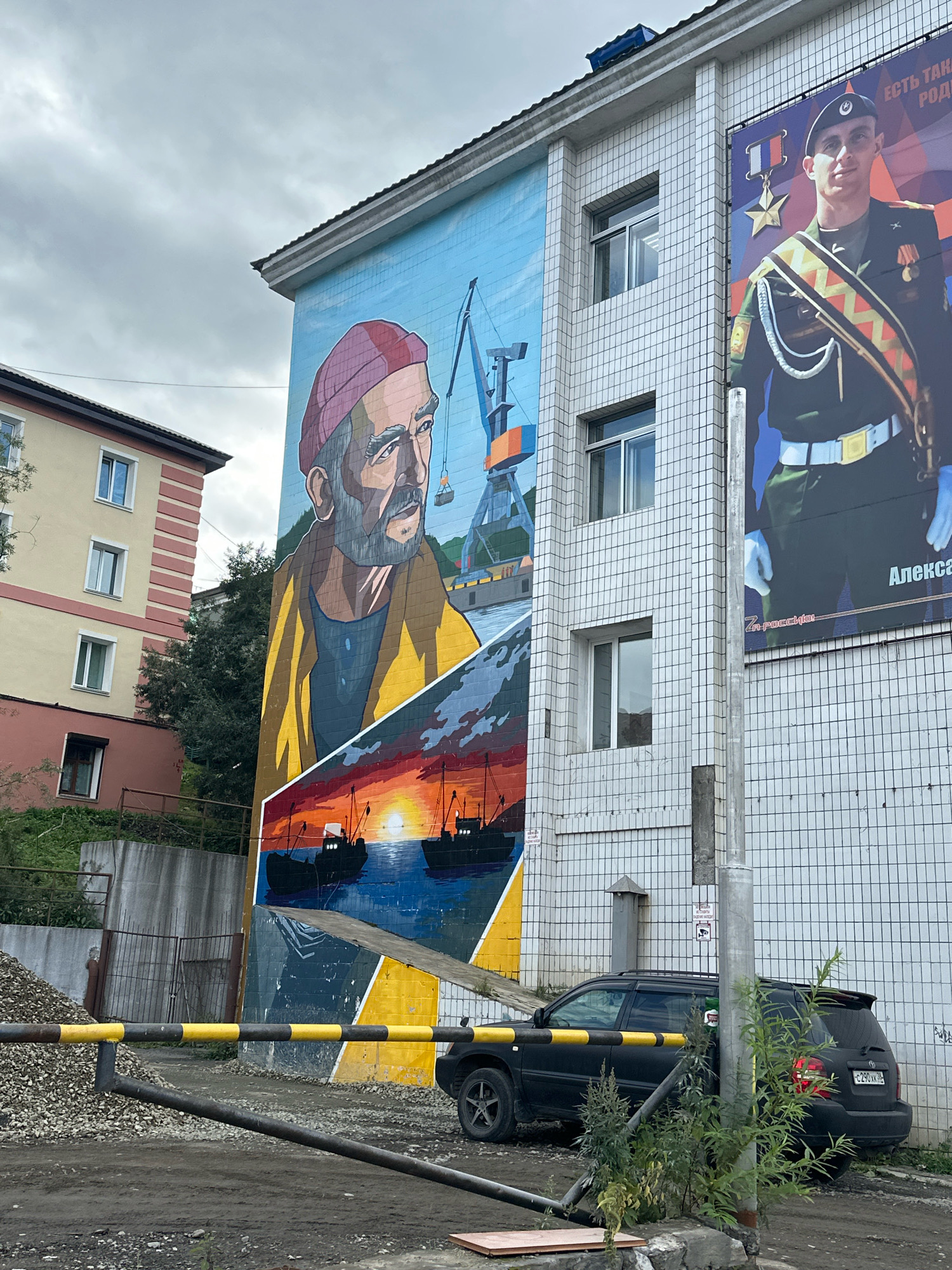 The image captures a mural on the side of a building in Petropavlovsk-Kamchatsky, Russia. The mural depicts a man with a beard wearing a red beanie and a yellow coat, looking out at a scene of boats on the water during sunset, with cranes in the background. To the right of the mural, a large poster features a man in a military uniform, identified as "Алекса...". The building itself has a tiled facade. In the foreground, a black SUV is parked, and a yellow and black striped barrier is in place. A pile of gravel and some vegetation are also visible. The sky is overcast.