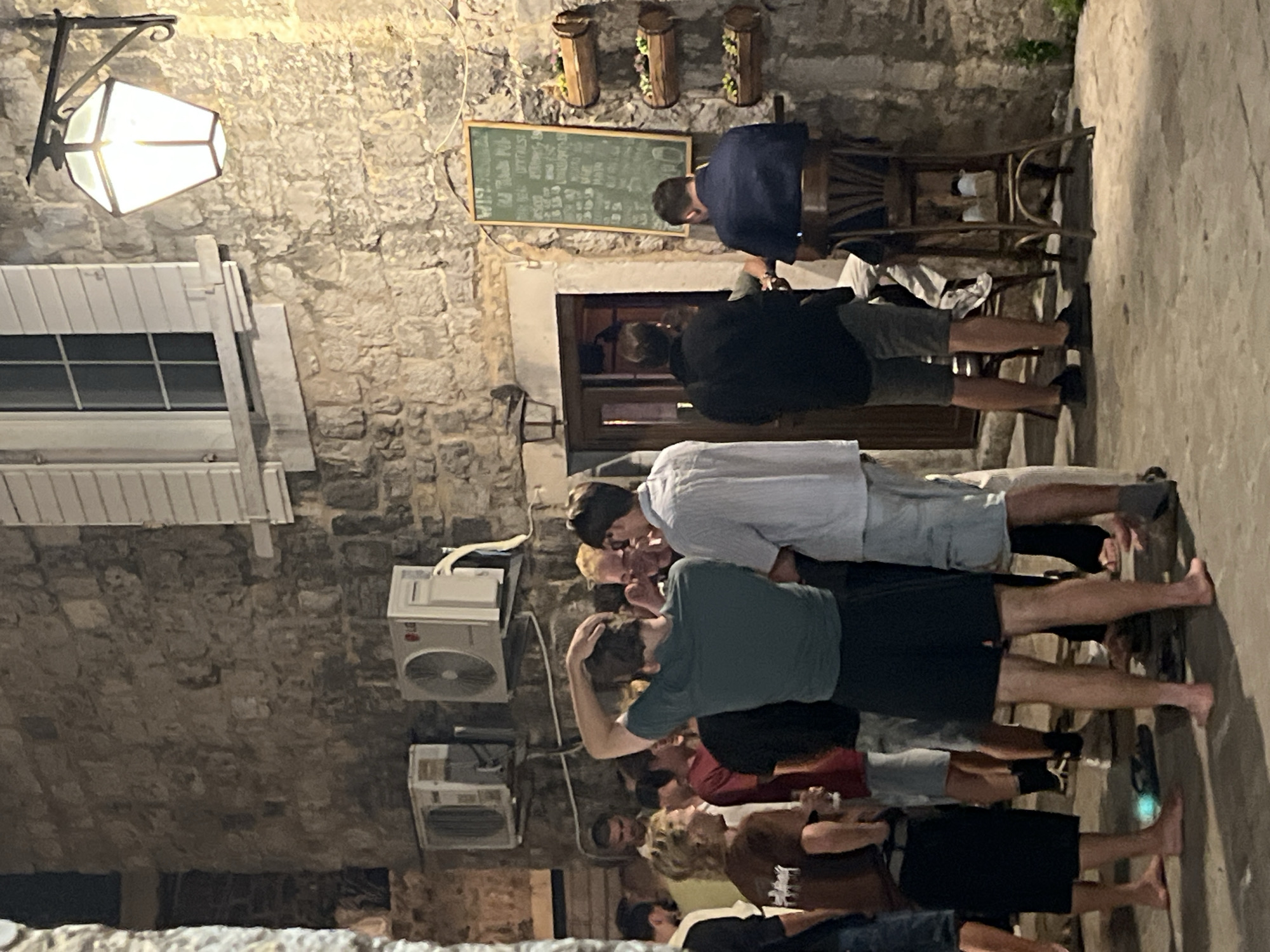 In the city of Budva, Montenegro, a group of people are gathered outside a building with a stone facade. The scene takes place at night, illuminated by a hexagonal lantern mounted on the wall and ambient light from the building's entrance. Several individuals, predominantly young men, are standing and conversing.
One man in a dark green t-shirt and black shorts is visible from the back, scratching his head. To his left, another man in a light blue and white striped shirt stands near a chalkboard sign that displays text, likely a menu or specials. To the right of the chalkboard, a man in a navy blue shirt is seated at a high stool.
In the foreground, several people are casually dressed, with some appearing to be barefoot on the cobblestone ground. The building features two large outdoor air conditioning units mounted on the stone wall. Wooden planters with flowers are attached to the wall above a doorway. The overall atmosphere suggests a relaxed social gathering outside a bar or restaurant.