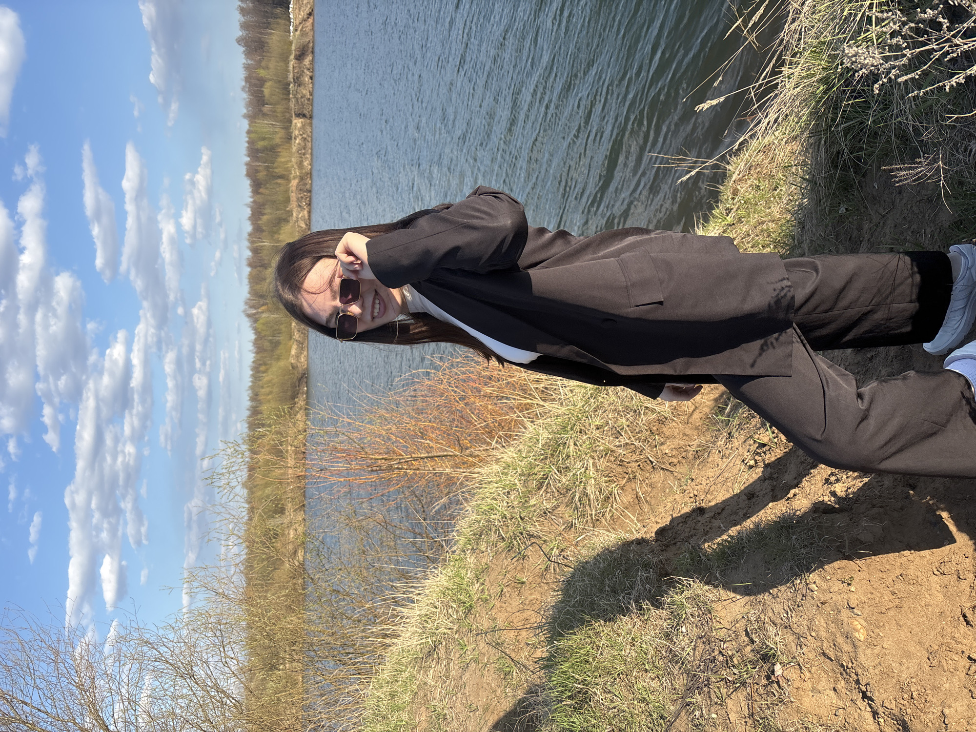 A young woman in a black suit and sunglasses stands on a grassy, dirt embankment near a body of water. Behind her is the riverbank and a forest. The sky is blue with scattered clouds. The photo was taken during the day in Zaosinovo, Russia.