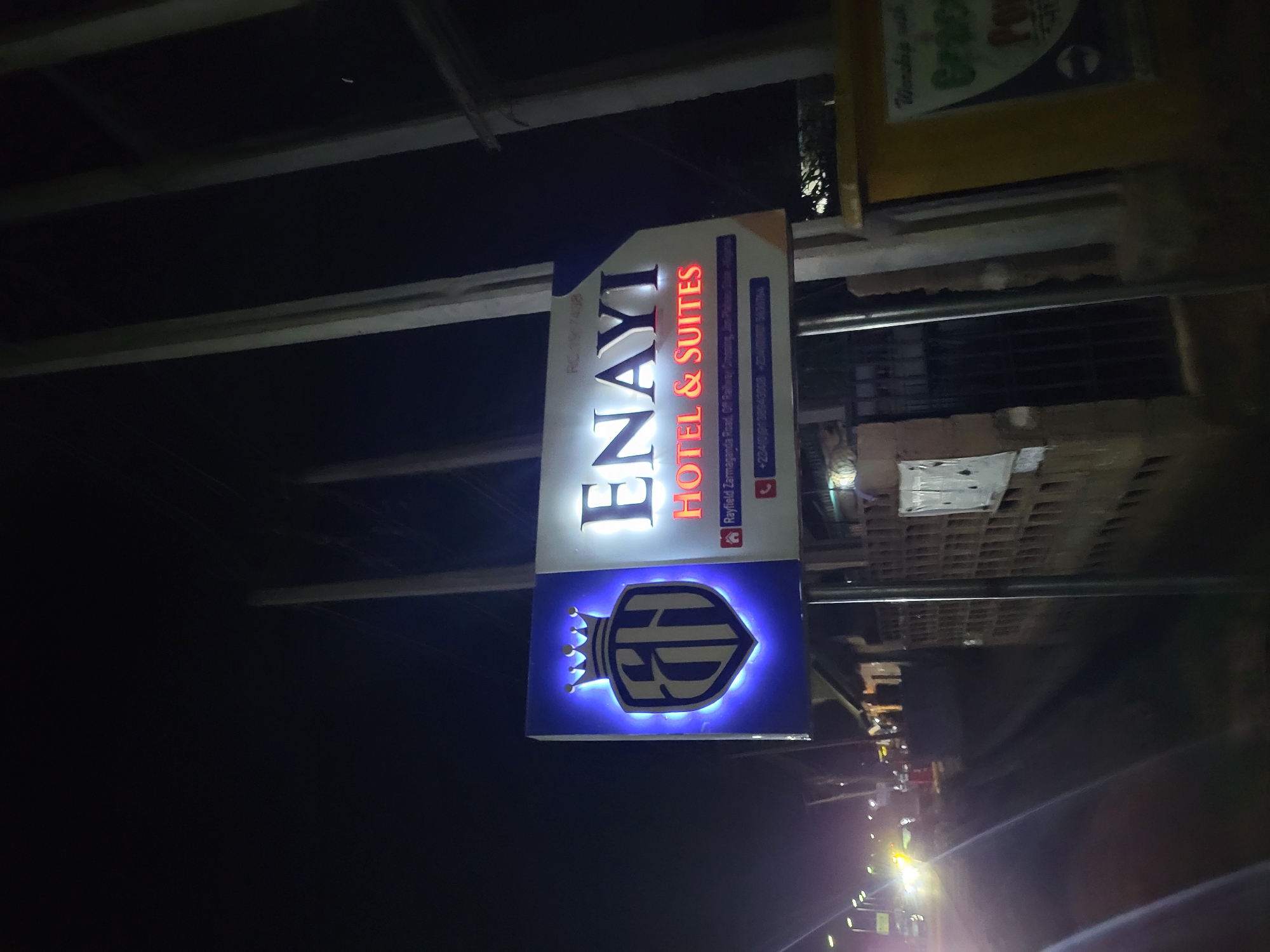 The image shows a sign for "ENAYI HOTEL & SUITES" located on Rayfield Zarmaganda Road, Off Railway, in an unknown city. The sign is illuminated and features the hotel's name, the phrase "HOTEL & SUITES," and the hotel's logo in a stylized shield shape with a crown above it. The logo is also illuminated. The sign also includes the registration number (RC 1907408), location, and phone number (+234(0)938943008). The background appears to be dark, suggesting that the photo was taken at night. Streetlights are visible in the distance.