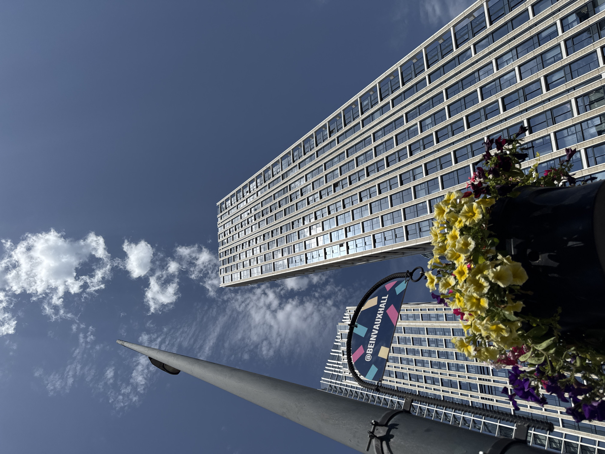 This low-angle shot captures a modern architectural scene against a bright blue sky with scattered white clouds. The primary subjects are two large, contemporary buildings with repeating patterns of windows. The building in the foreground, on the right, is partially obscured by a hanging planter filled with yellow and purple flowers. Behind this building, another similar structure stretches across the frame.
A tall, grey lamppost is prominently featured in the lower left portion of the image, angling upwards. Attached to the lamppost, a circular banner is visible, displaying the text "@BEINVAUXHALL" in white letters against a dark blue background, interspersed with colorful geometric shapes of pink, teal, and yellow.
The scene suggests an urban environment, likely a public space given the lamppost and banner. The bright sky and the sunlight reflecting off the windows indicate it is daytime, possibly late morning or early afternoon, with fair weather. The perspective, looking up from ground level, emphasizes the scale and modernity of the architecture. The presence of flowers adds a touch of nature and color to the otherwise geometric and man-made surroundings. There are no people visible in the image. The overall mood is one of urban dynamism and architectural presence. The location is indicated by the text on the banner to be Vauxhall.