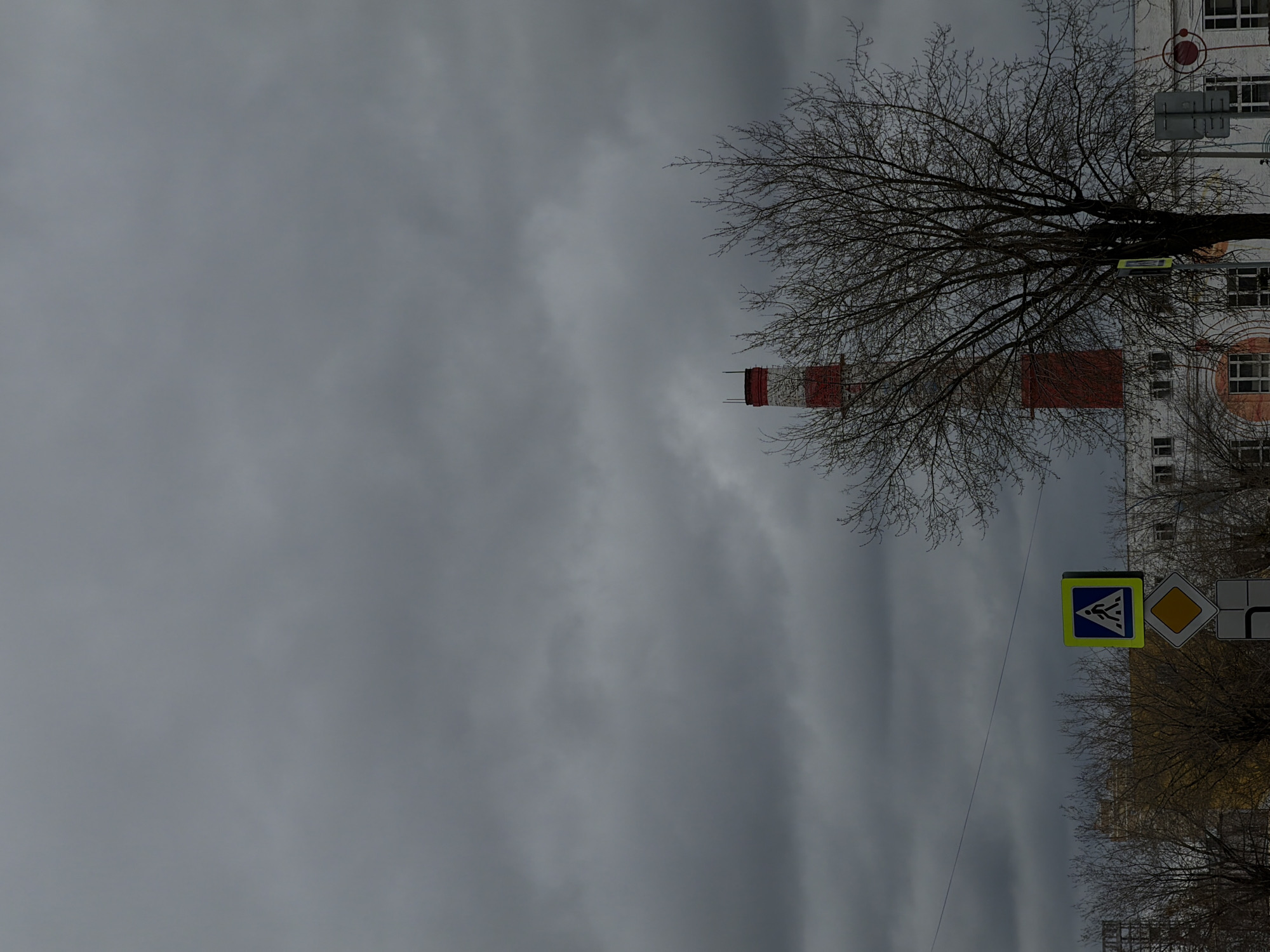 The image shows a street scene in Perm, Russia on an overcast day. A tall, striped chimney stands out against the gray sky. A bare tree obscures a white building. Road signs are visible, including a pedestrian crossing sign.