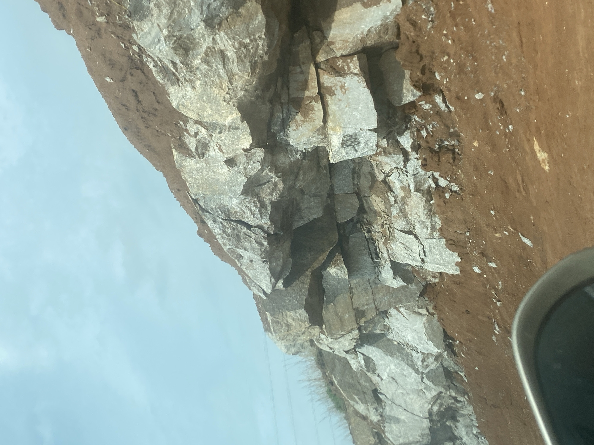 The image displays a rugged rocky cliff face, viewed from a perspective that suggests it was taken from a vehicle. The cliff, composed of large, fractured grey and off-white rocks, dominates the upper part of the scene. Above the main rock formation, a distinct layer of reddish-brown earth or soil is visible, scattered with small white fragments. Below the cliff, a wide expanse of bright, clear blue sky indicates daytime conditions.
In the bottom right corner, a partial view of a curved, grey metallic object, likely part of a vehicle's interior like a dashboard or rearview mirror housing, is visible. This detail suggests the photograph was captured while moving through the landscape of Gosa, Nigeria. Thin, dark lines, possibly power lines or similar cables, can be seen against the sky, stretching horizontally across the lower-middle section of the image.
Notable details on the rock face include numerous cracks and fissures, highlighting its weathered texture. Near the center of the rock formation, on a lighter-colored section, black markings are present. These appear to be graffiti or symbols; one distinct mark resembles a stylized number "2" or "Z," possibly followed by other indistinct characters, or an "S" with an arrow. The scene captures a natural geological feature alongside human elements, providing a glimpse of the environment in Gosa.
