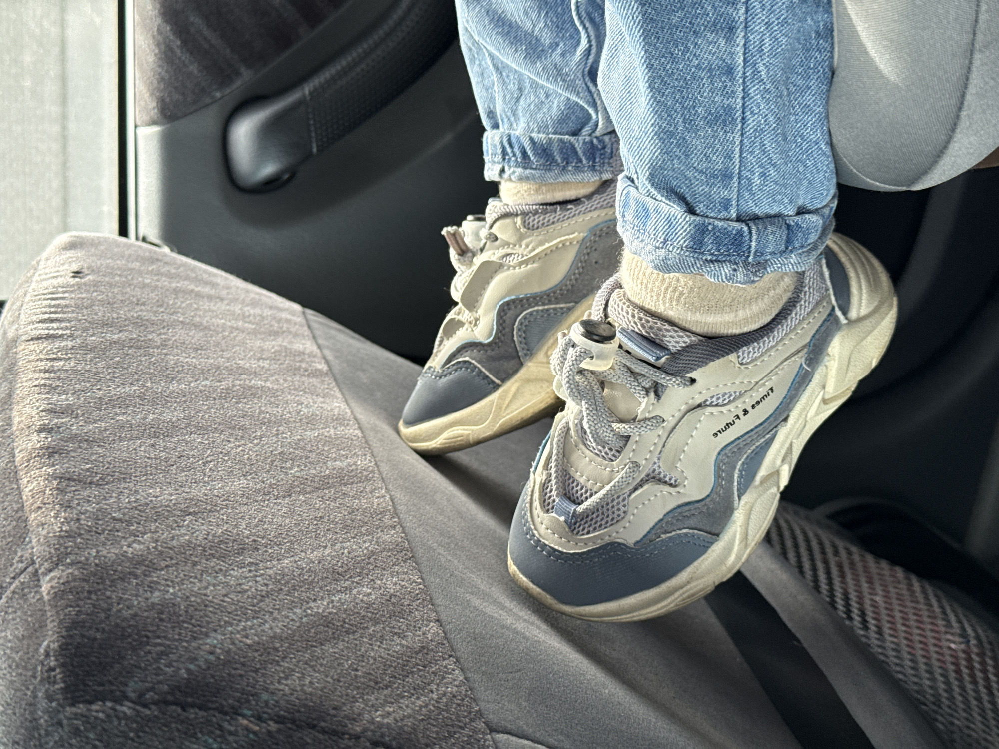 The image shows a child's legs, clad in light blue jeans cuffed at the ankles and off-white socks, sitting on a grey cloth-covered car seat. The child is wearing two-toned sneakers - light grey and blue. The sneakers have white soles and grey laces. The car's interior is visible with a dark grey door panel in the background. The scene depicts a casual, static setting. The location is assumed to be in a car, possibly in Krasnoyarsk, Russia, without any visual confirmation.